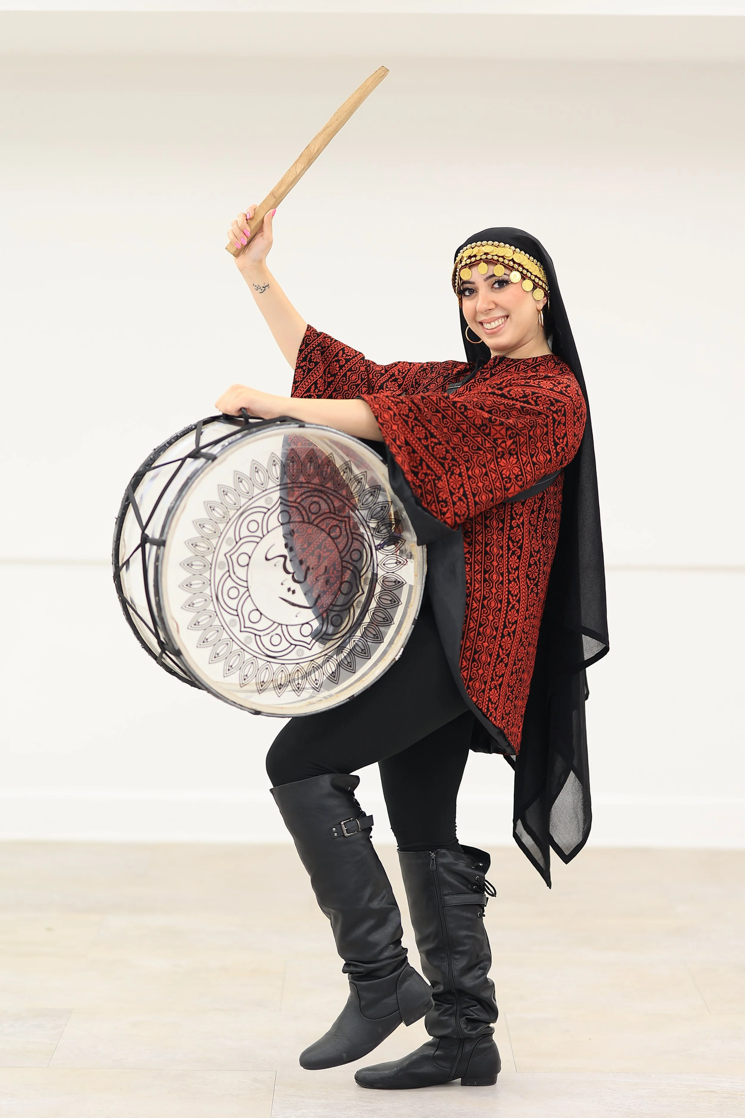 A woman in traditional Palestinian clothing with gold decorative headpiece is playing a large drum with a wooden stick. She is smiling and standing on one leg, wearing black boots.