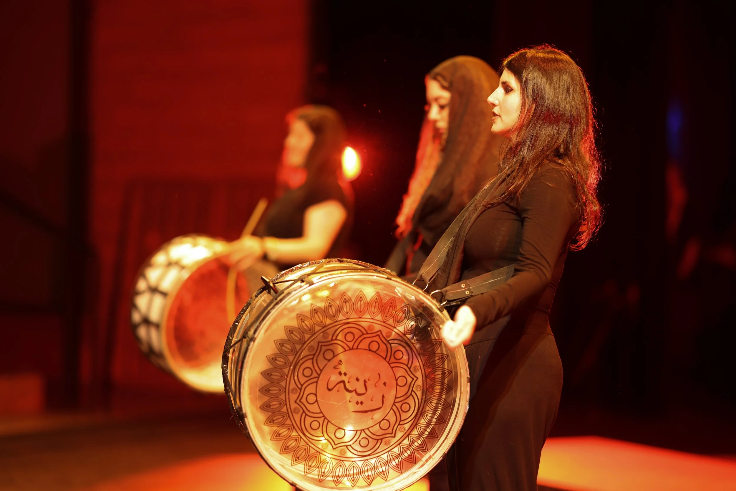 Three women are playing large hand drums on stage, illuminated by warm stage lights. Their focus is on their drums during a performance.