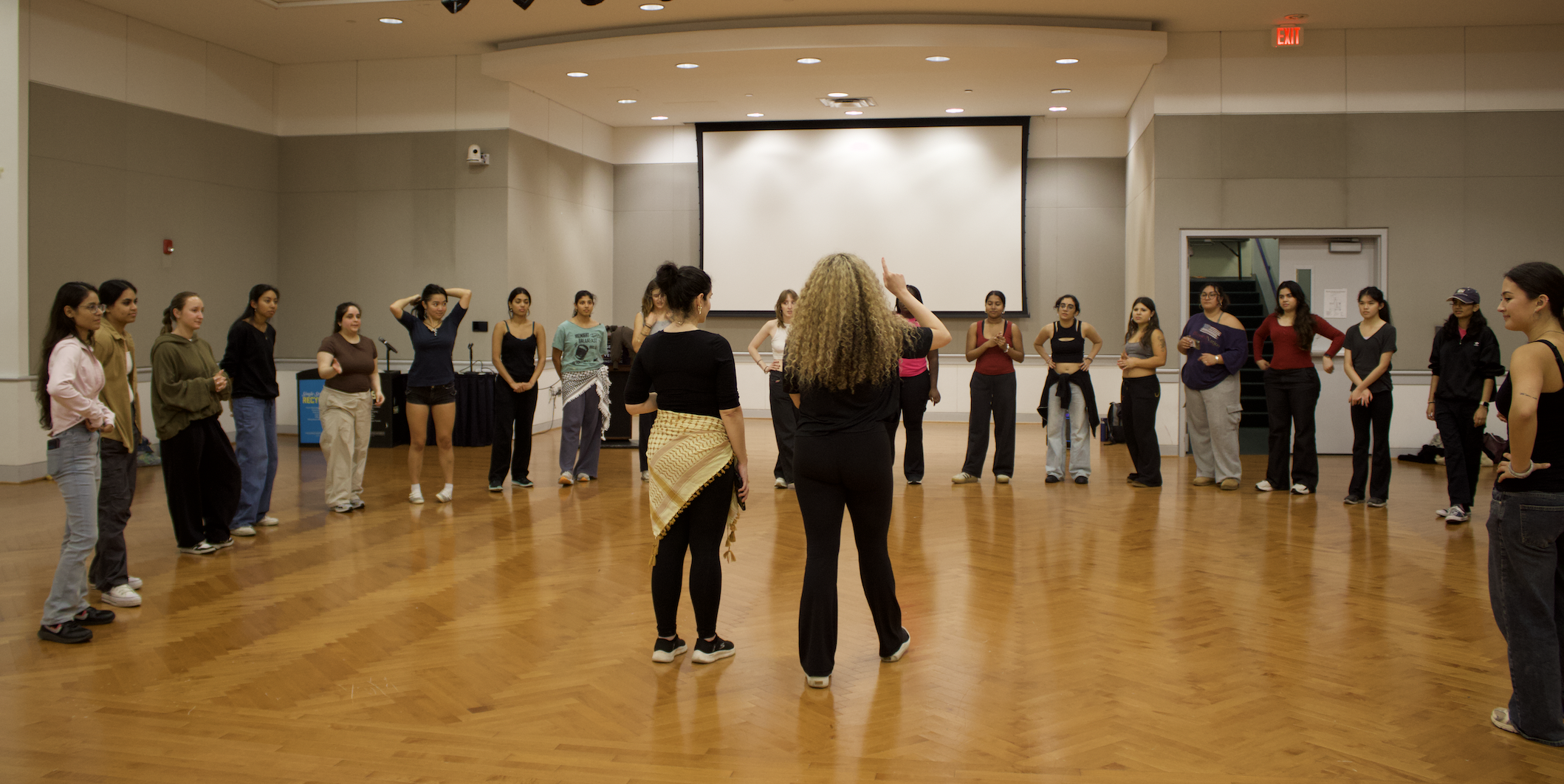 A group of women standing and listening to a woman with curly blonde hair, in a circle in a spacious room with wooden floors, a large projection screen, and a staircase in the background.