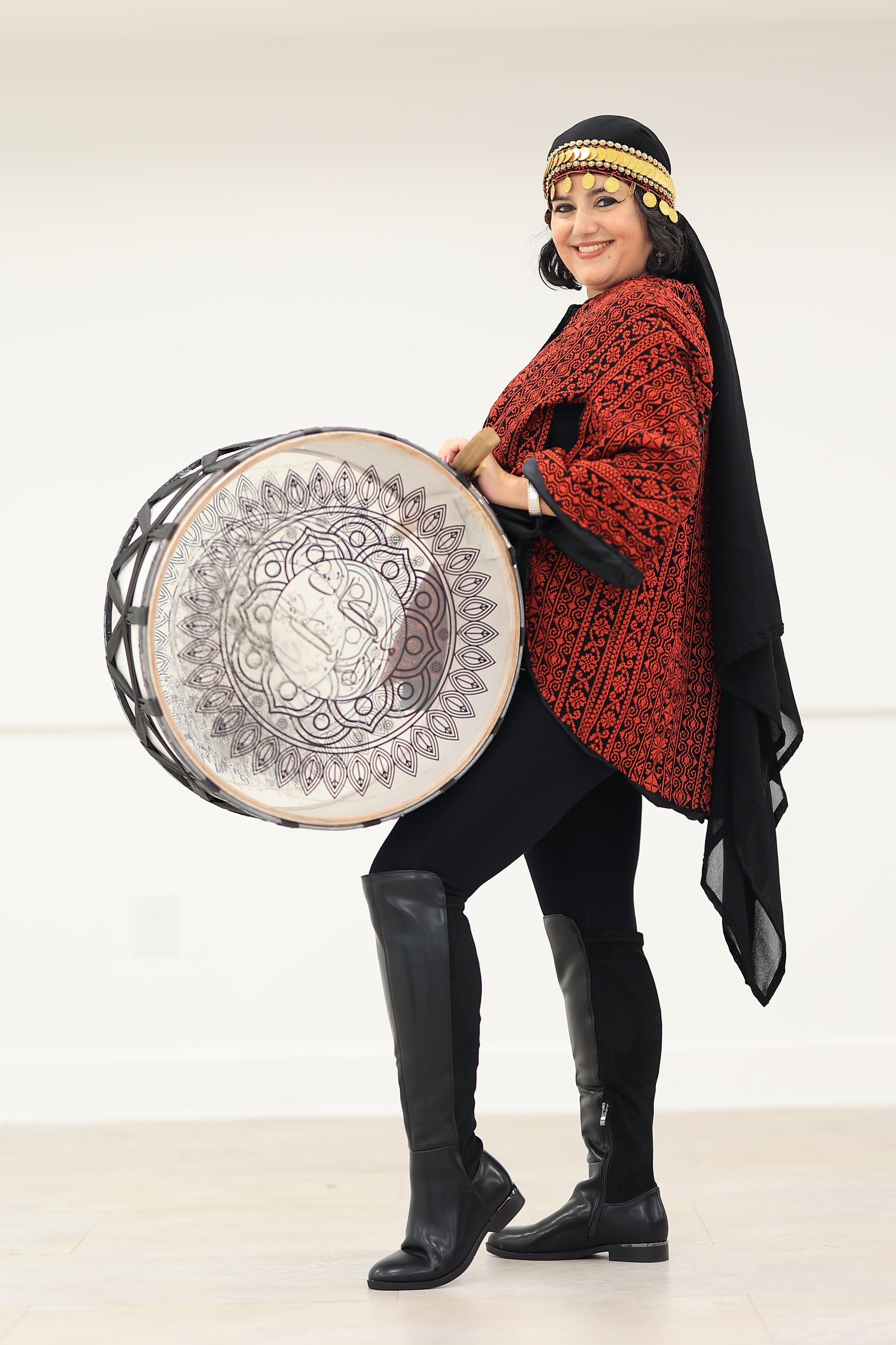 A woman in traditional Palestinian clothing with gold decorative headpiece is playing a large drum with a wooden stick. She is smiling and standing on one leg, wearing black boots.