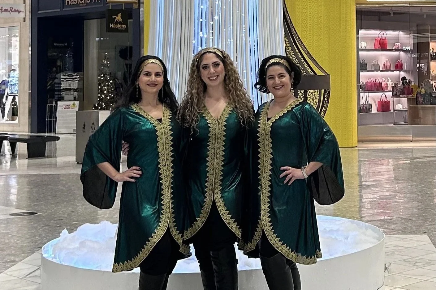 Three women in matching green and gold traditional dresses standing inside a shopping mall in front of a white decorative float.