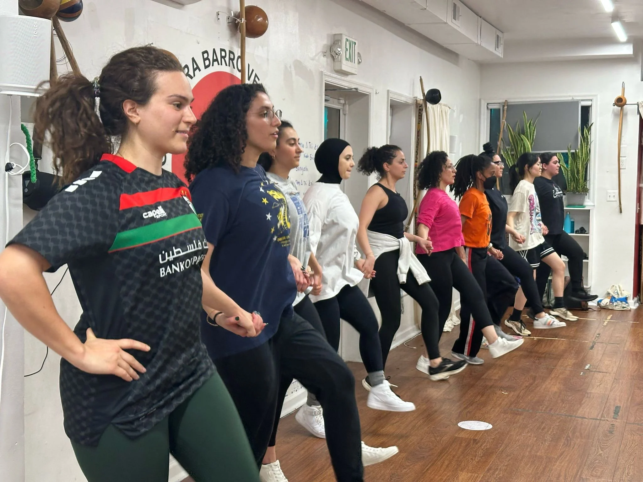 A group of women in a dabke dance class practicing synchronized choreography, standing side by side with hands on their hips and one leg lifted, in a well-lit room with white walls and wooden flooring.