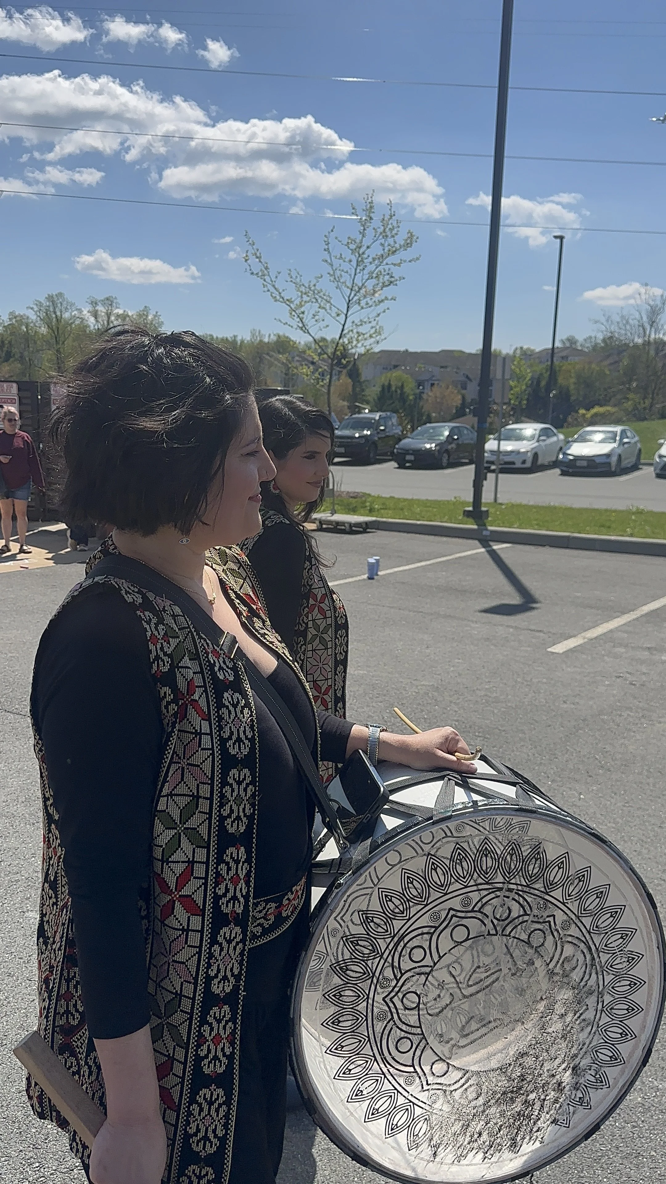 Two women standing outdoors on a sunny day, one playing a large decorative drum, in a parking lot with cars, trees, and a blue sky with clouds.