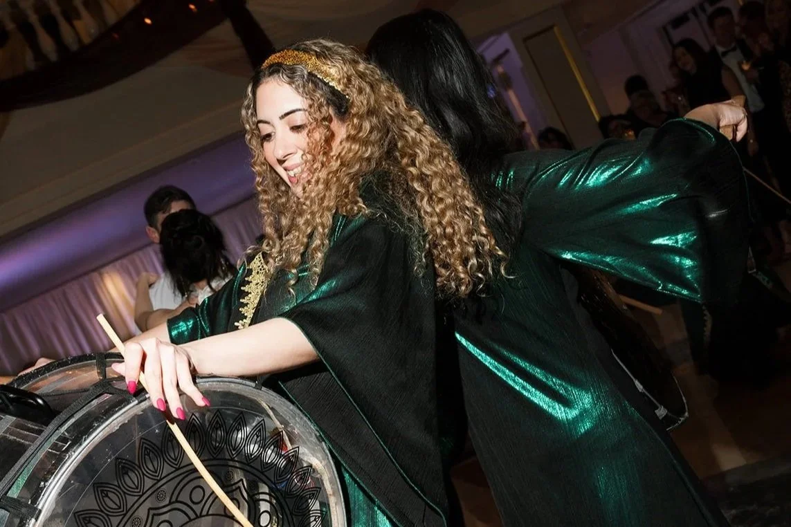 Two women dancing at a party, one with curly blonde hair and a black headband, smiling, and the other with straight black hair, wearing shiny green and black clothing, with a crowd of people in the background.