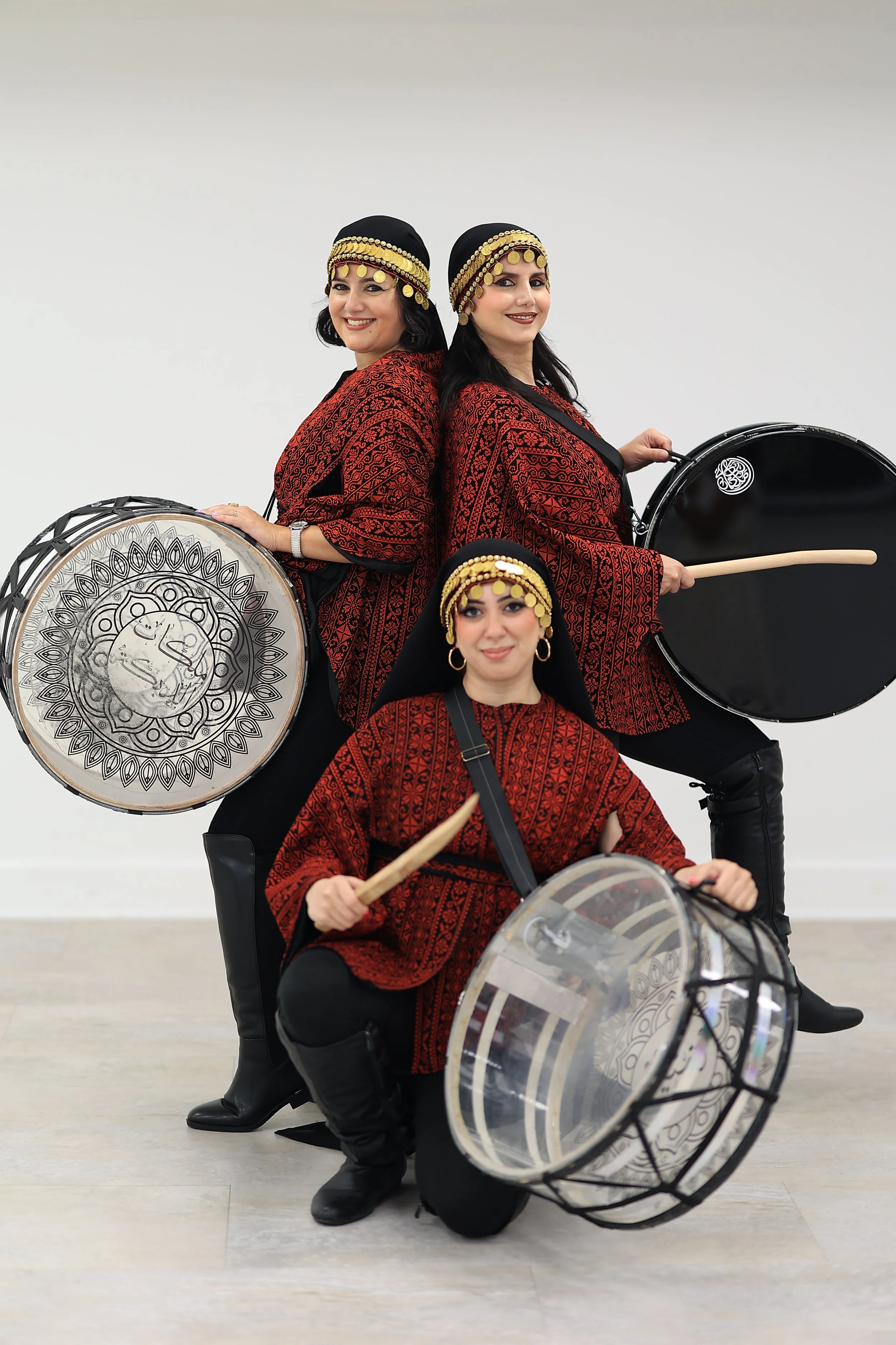 Three women dressed in Palestinian clothing with gold coin headpieces, holding drums, posing against a plain white background.