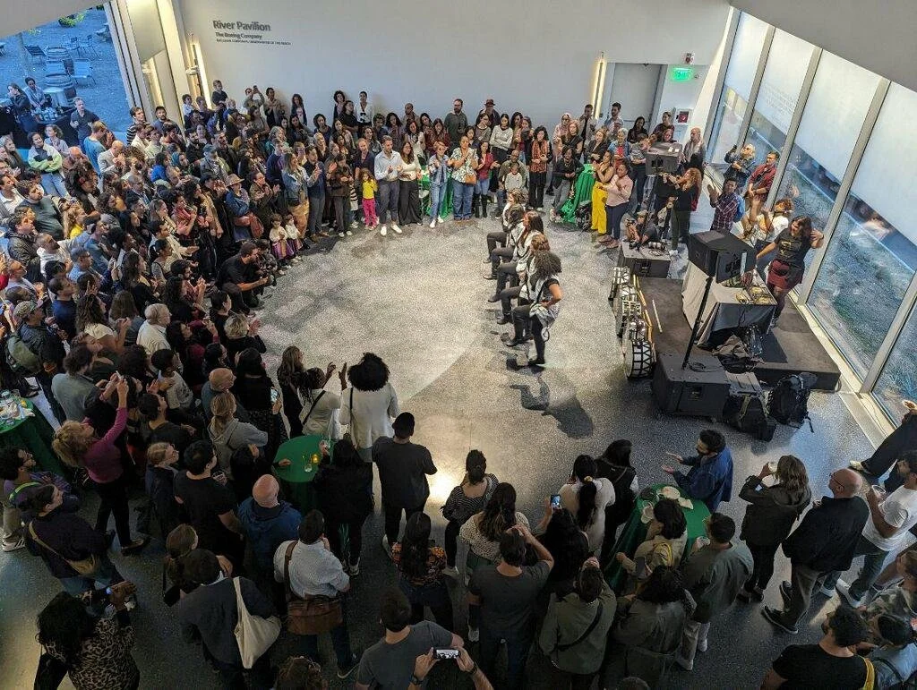 A large crowd of people gathered in a spacious, modern indoor venue with large floor-to-ceiling windows. Four people are seated in the center, performing or speaking, while the audience watches. There is a stage with sound equipment on the right side.