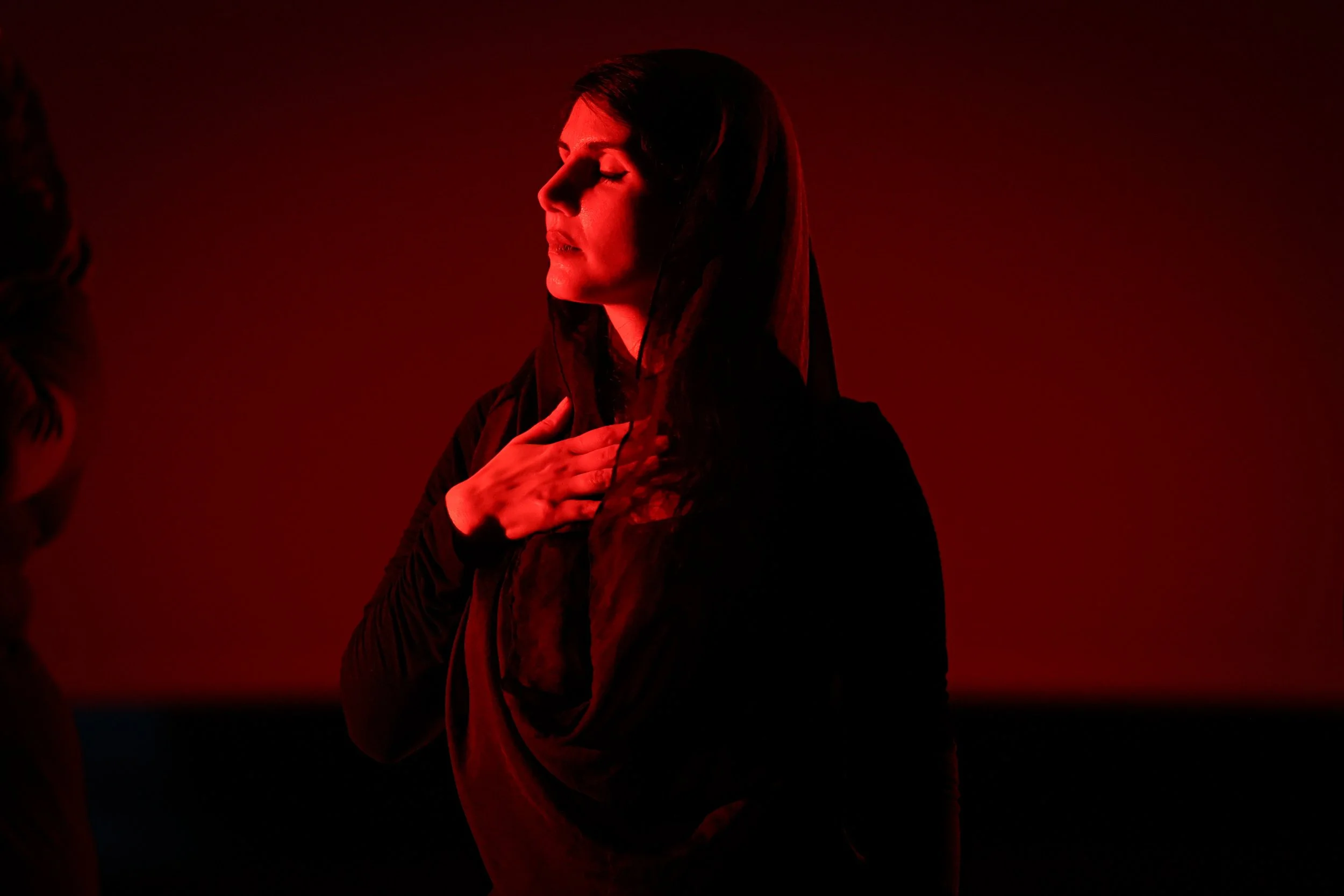 A woman with long dark hair and closed eyes, wearing dark clothing and a veil, holding her chest in front of a red background.