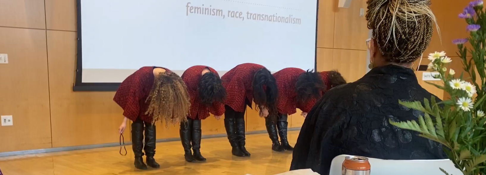 Four women bowing on stage, dressed in matching red outfits with black boots, during a performance or presentation. An audience member with braided hair and glasses is seated in front, with a flower arrangement and a soda can on the table nearby. A large screen behind displays text that reads 'feminism, race, transnationalism'.