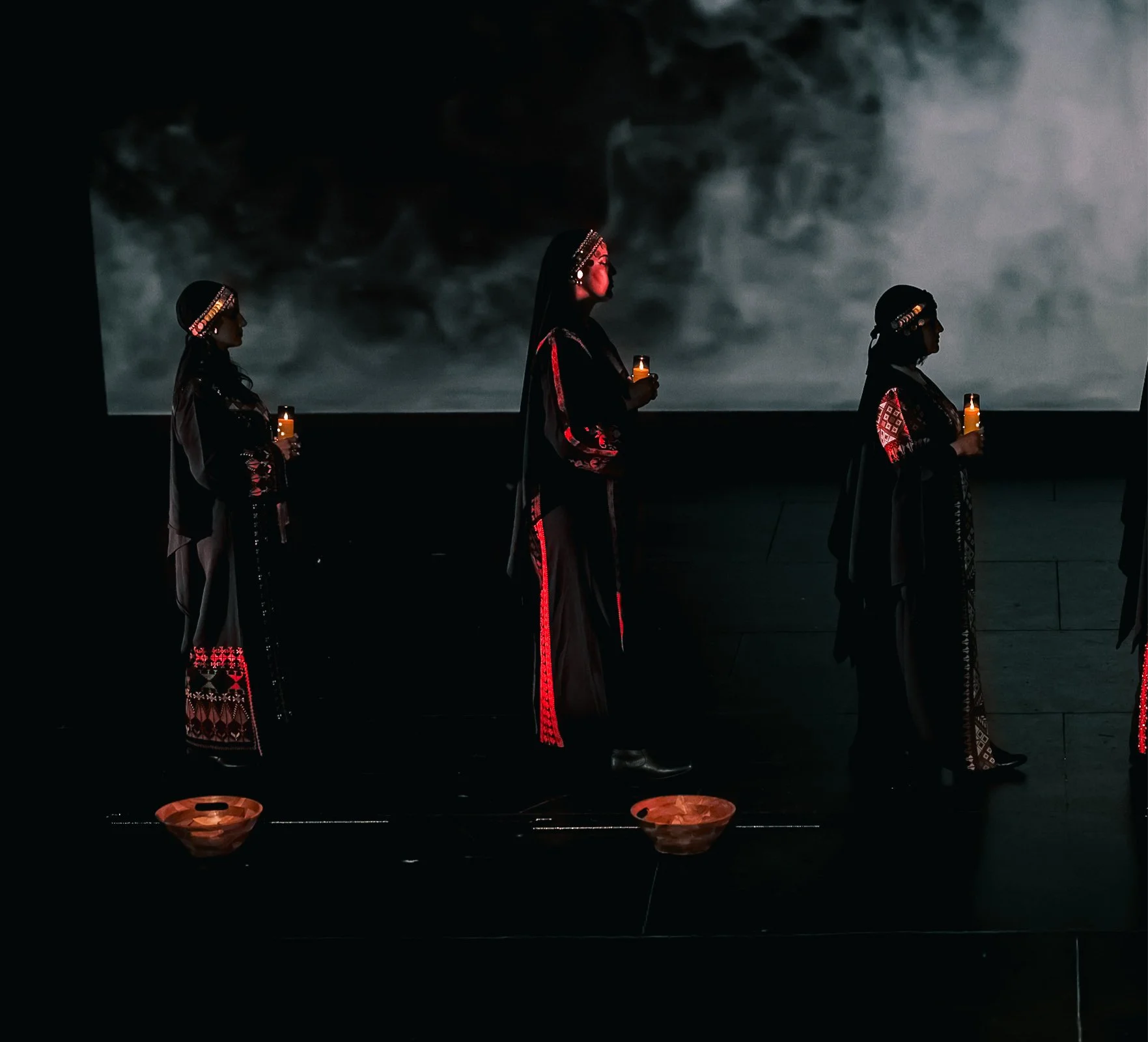 Three women wearing traditional embroidered clothing and headbands holding candles in a dark setting, with smoke projected in the background.