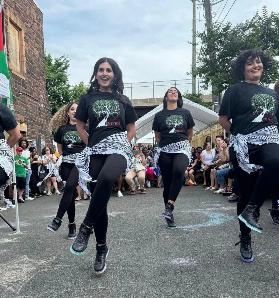 Four women wearing black T-shirts with a white tree design and the word 'Palestine', black pants, and black sneakers dance in a street during a gathering, with a crowd of spectators seated and standing behind them, and a clear sky above.