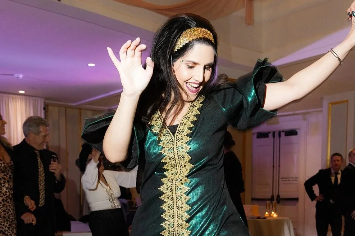 Woman dancing and smiling at an indoor party, wearing a shiny black dress with gold embroidery, gold headband, and black nail polish, with other guests in the background.