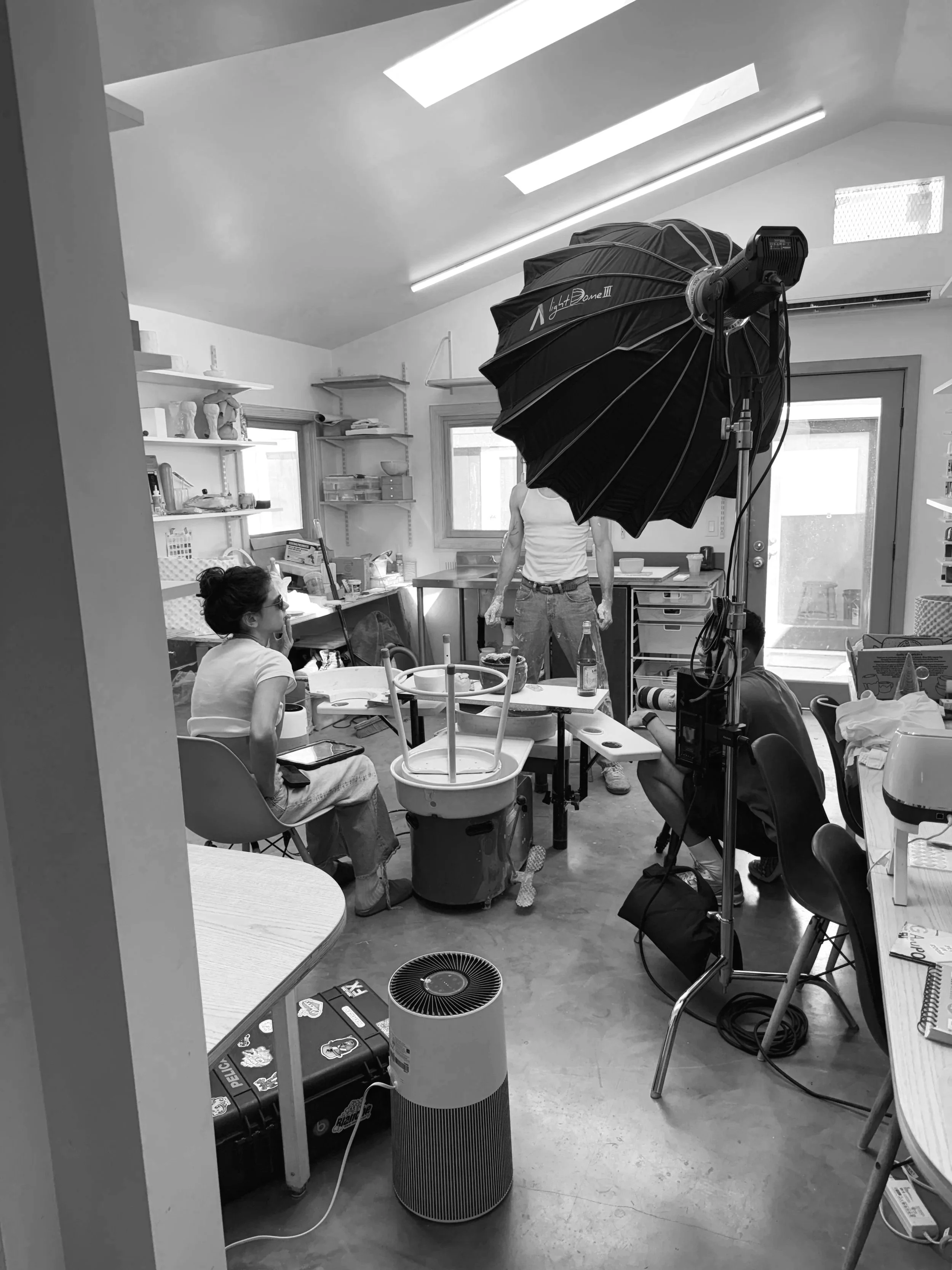 A photography studio with a large professional light, a person in a white tank top standing, a woman sitting on a chair, and various studio equipment, shelves, and windows in the background.