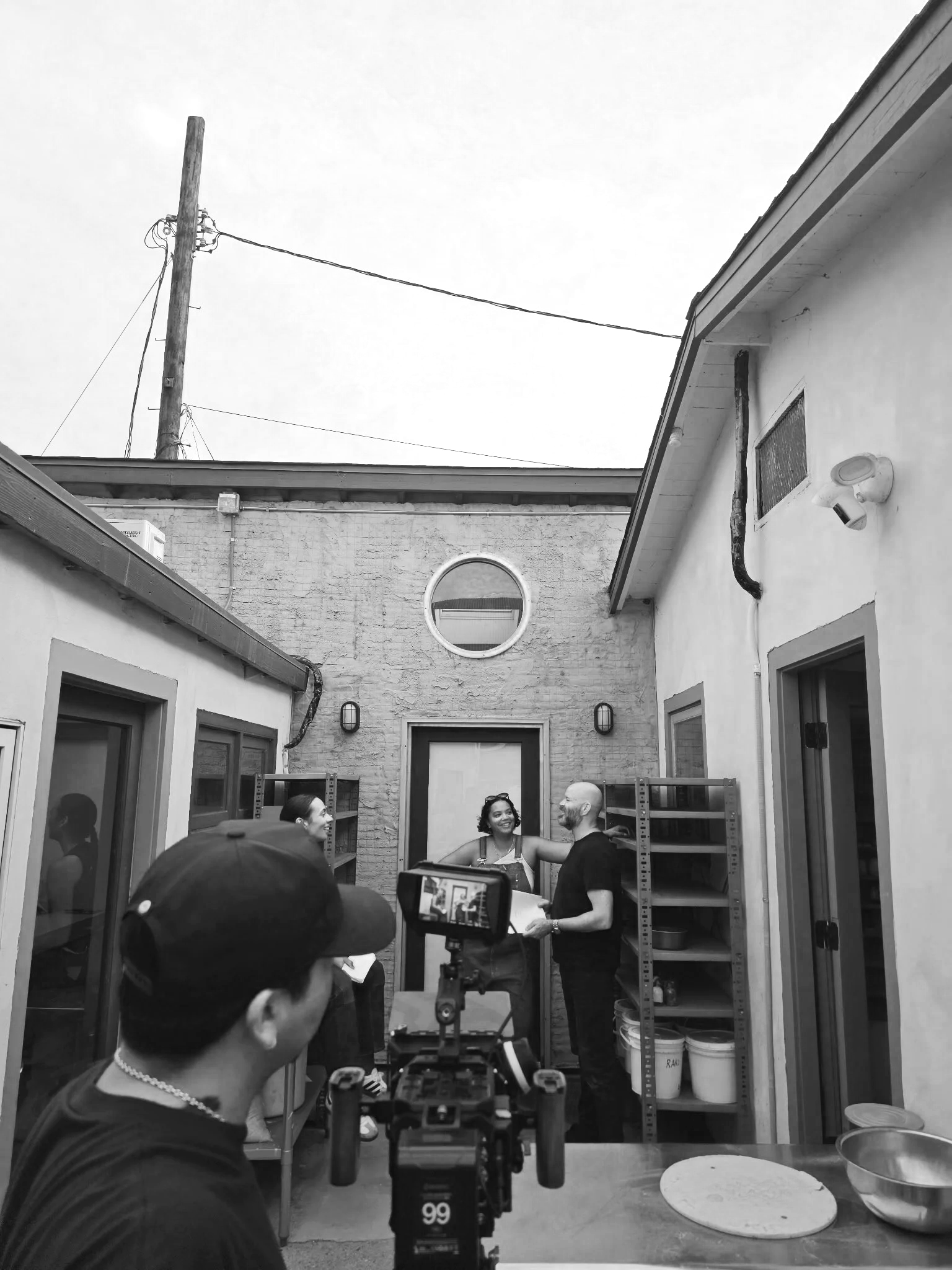 People having a conversation in a small urban courtyard, with a camera setup in the foreground capturing the scene, surrounded by shelves and wall fixtures.