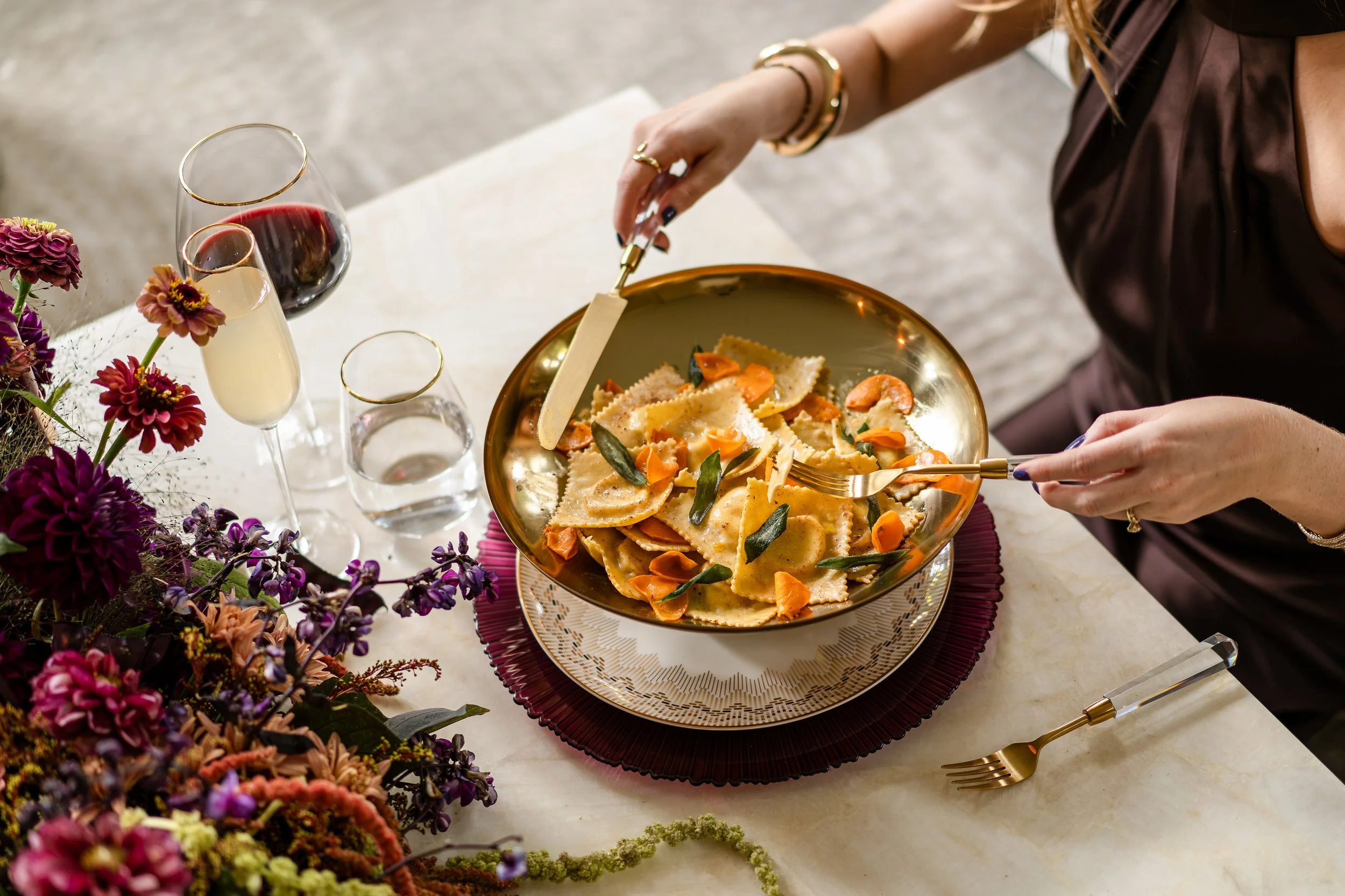 A person with long hair and jewelry serving a bowl of ravioli pasta with vegetables at a decorated table with flowers and wine.