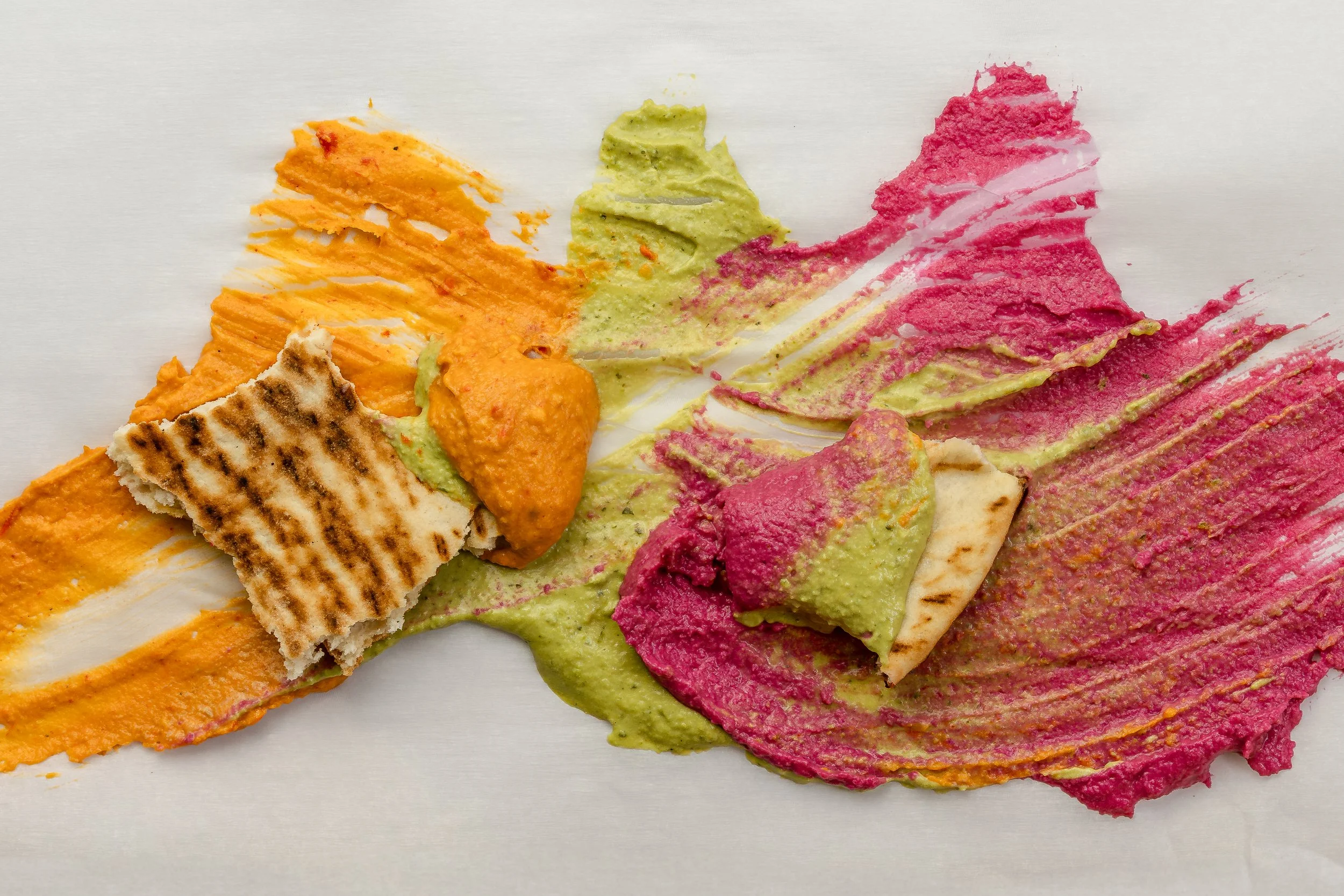 Colorful smeared hummus on a white surface with some grill marks on a piece of flatbread.