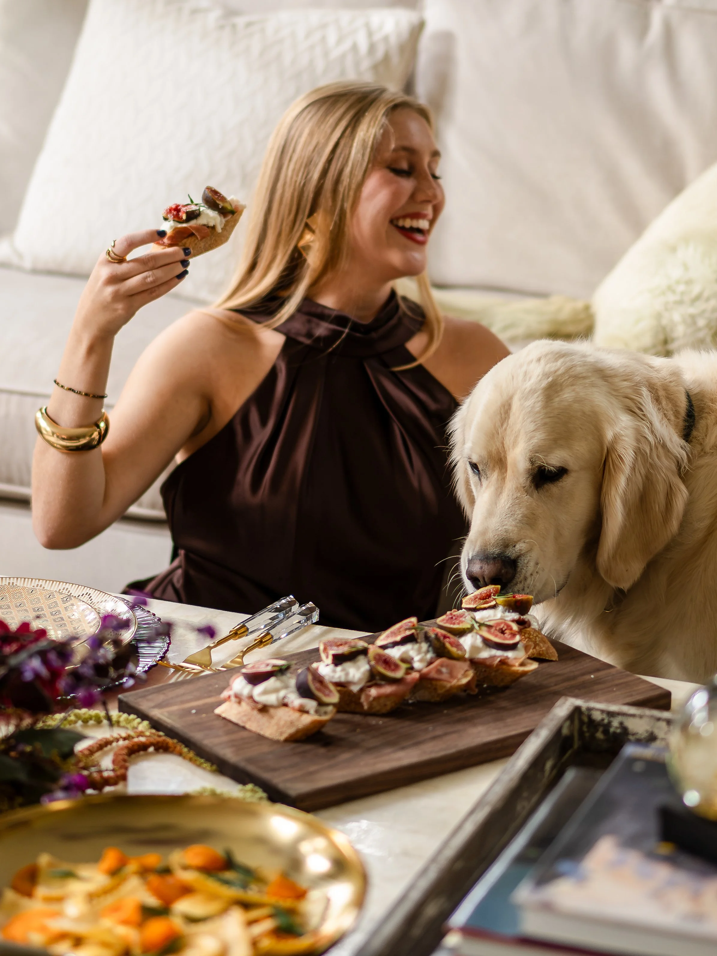 A woman smiling while holding a piece of bread with toppings, sitting at a table with various open-faced sandwiches and food items, and a dog licking a piece of bread.