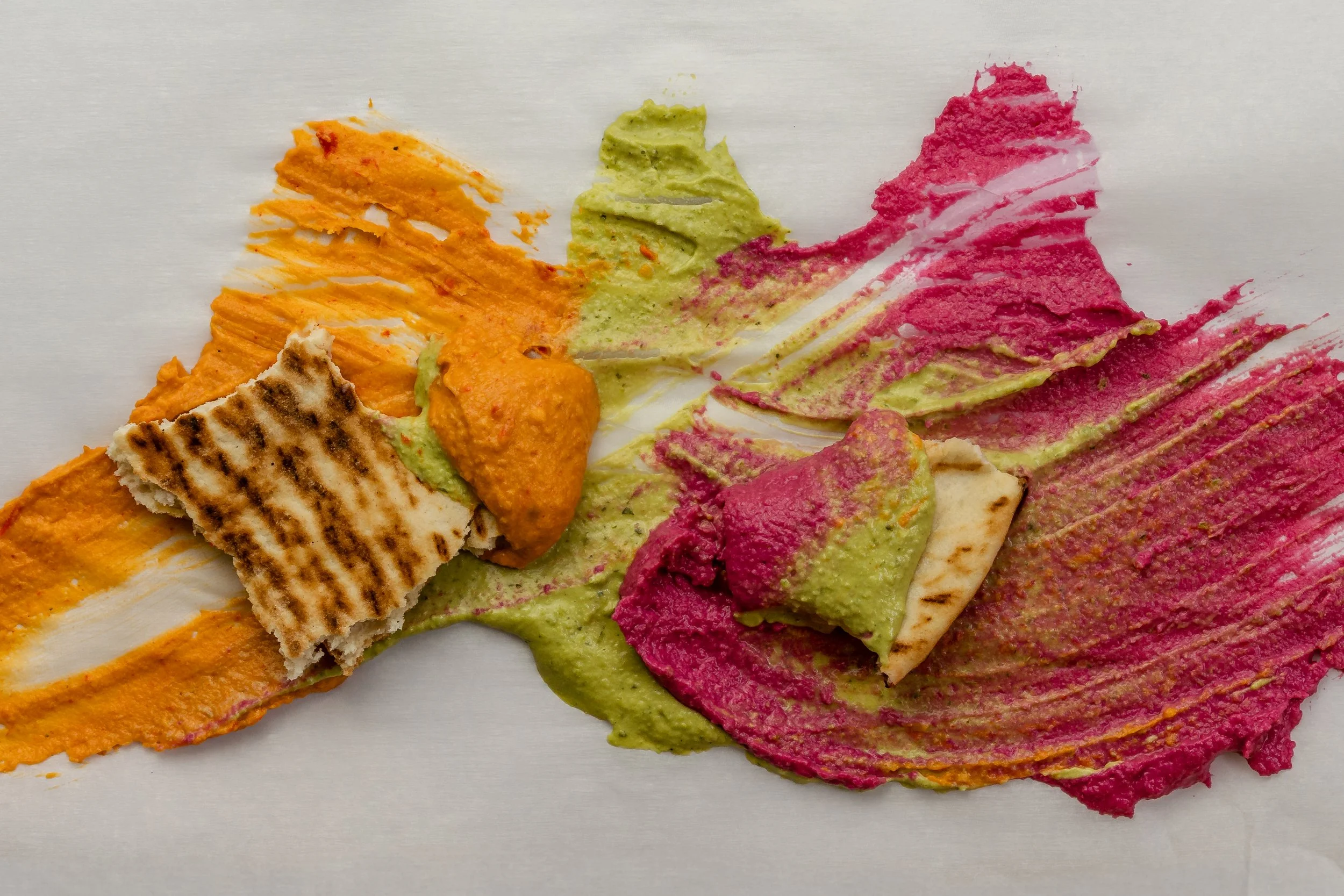 Colorful smeared hummus on white surface with grilled flatbread pieces.