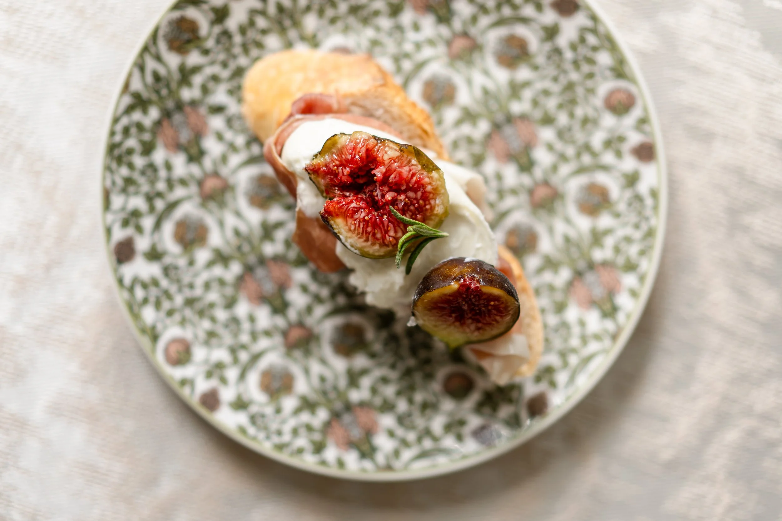 Close-up of an open-faced sandwich with figs, cheese, prosciutto, and herbs on a patterned plate.