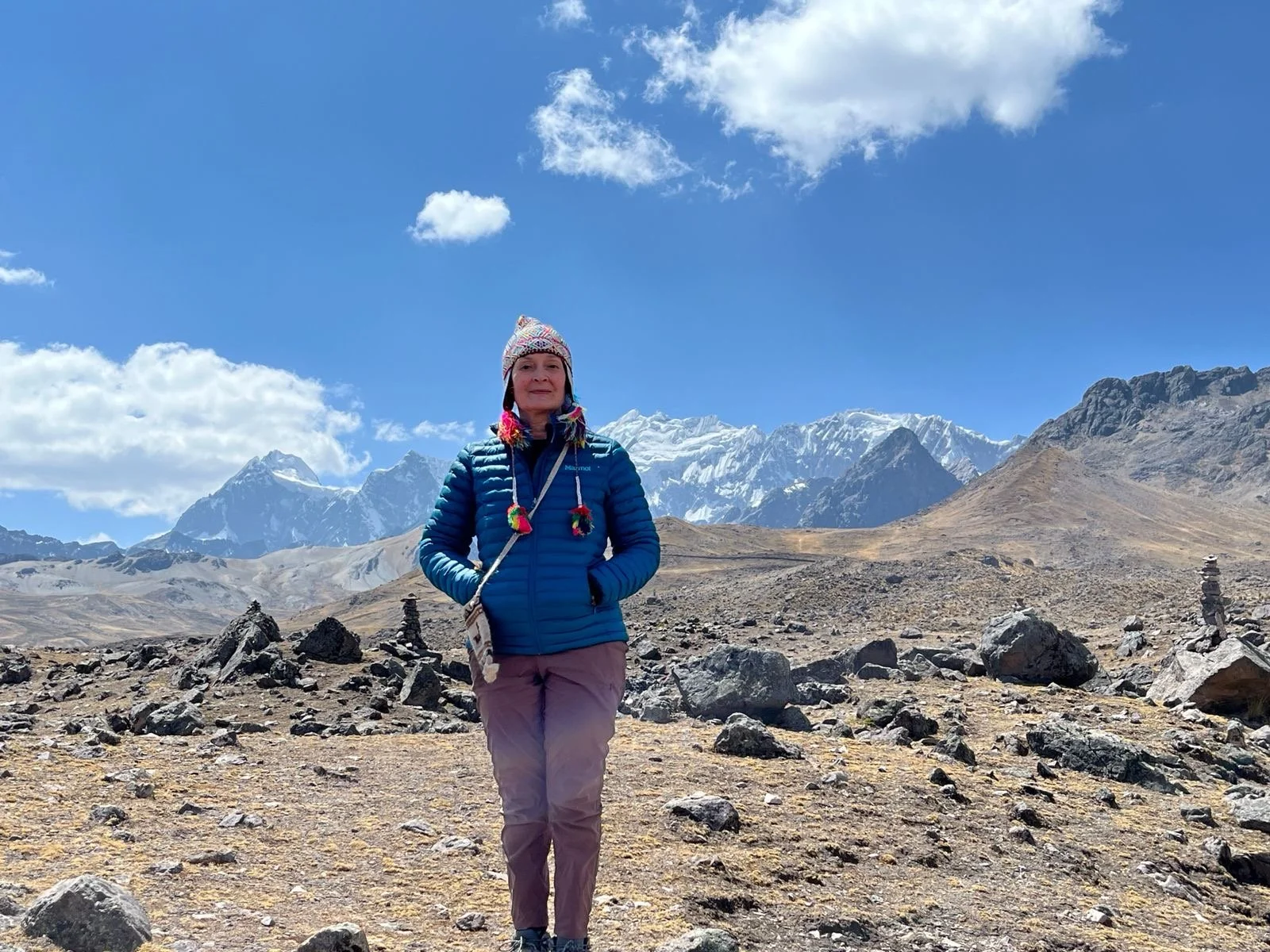 A woman standing outdoors in Peru with snow-capped peaks, wearing a blue jacket, purple pants, and a colorful knit hat with pom-poms.