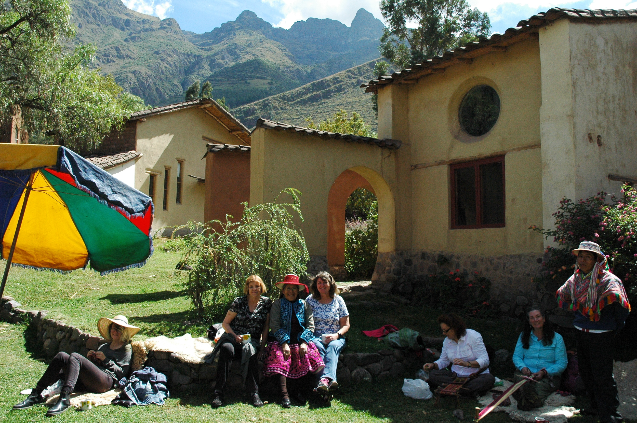 Group of women sitting on a stone wall and on the grass outside a yellow house with a circular window, in a lush mountainous landscape under a blue sky.