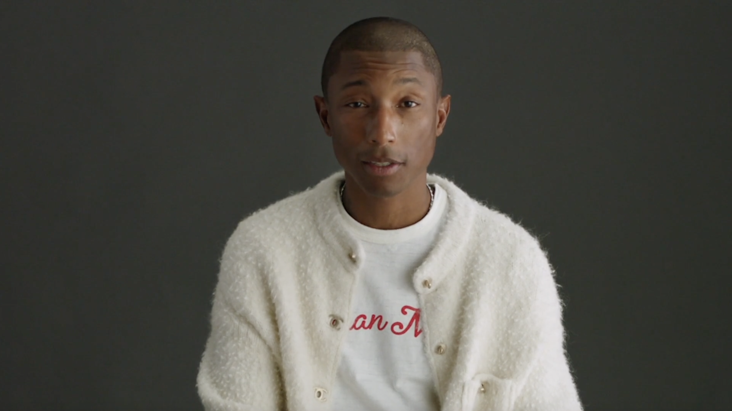 A young man with short hair, wearing a white textured cardigan over a white t-shirt with red text, sits against a dark gray background.