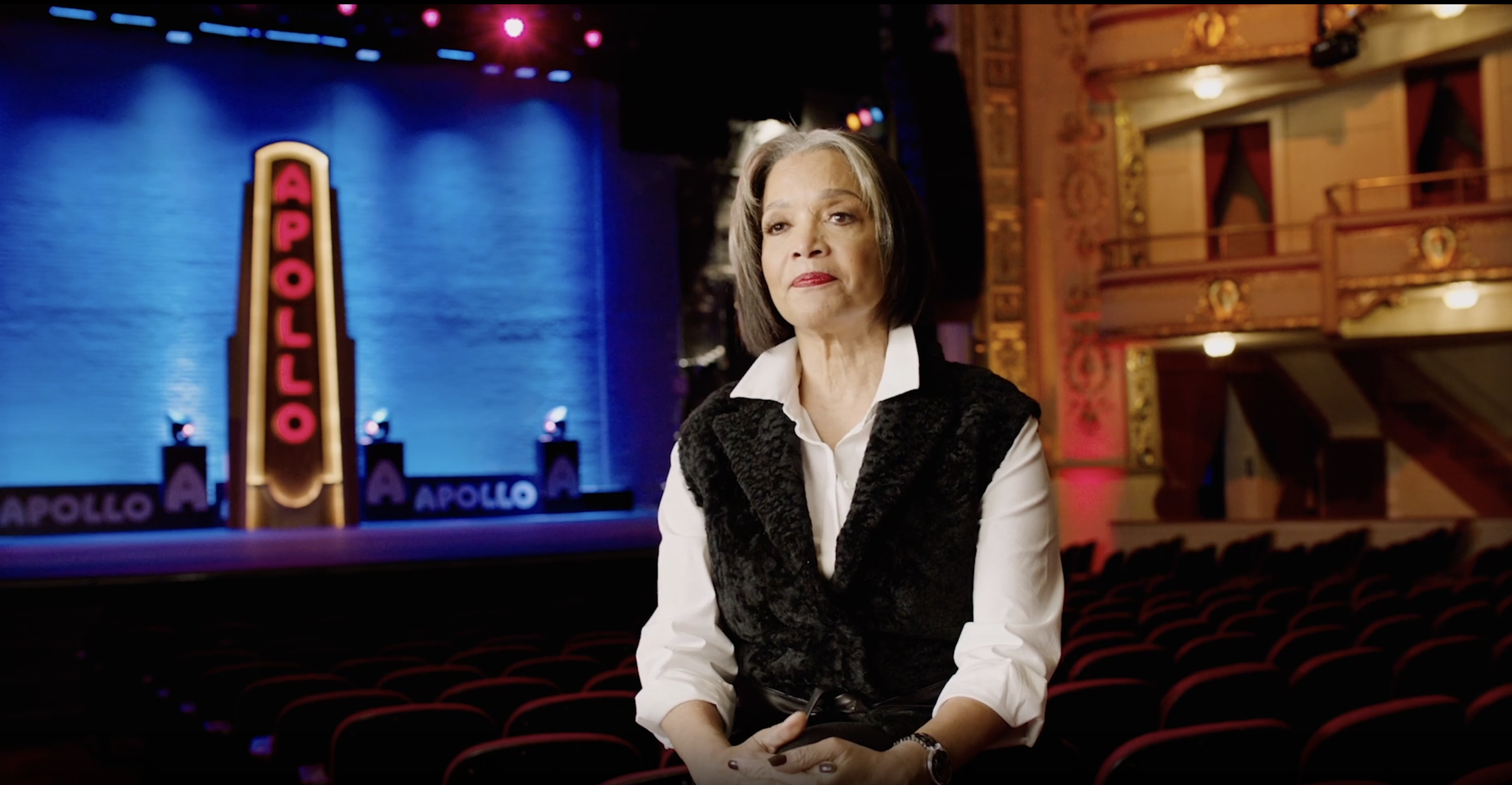 An older woman sitting in an empty theater, with a stage in the background displaying a neon sign that reads 'APOLLO' and theater seating.