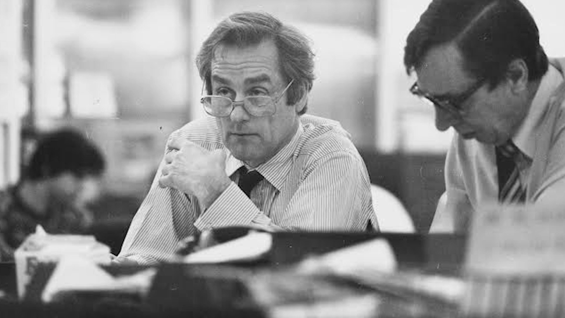 A black and white photo of two men in a meeting or office setting. The man on the left has glasses, gray hair, and is wearing a striped shirt with a tie. He looks towards the camera with a serious expression. The man on the right has dark hair, glasses, and is wearing a light-colored shirt with a tie. He appears to be speaking or explaining something, leaning slightly forward.