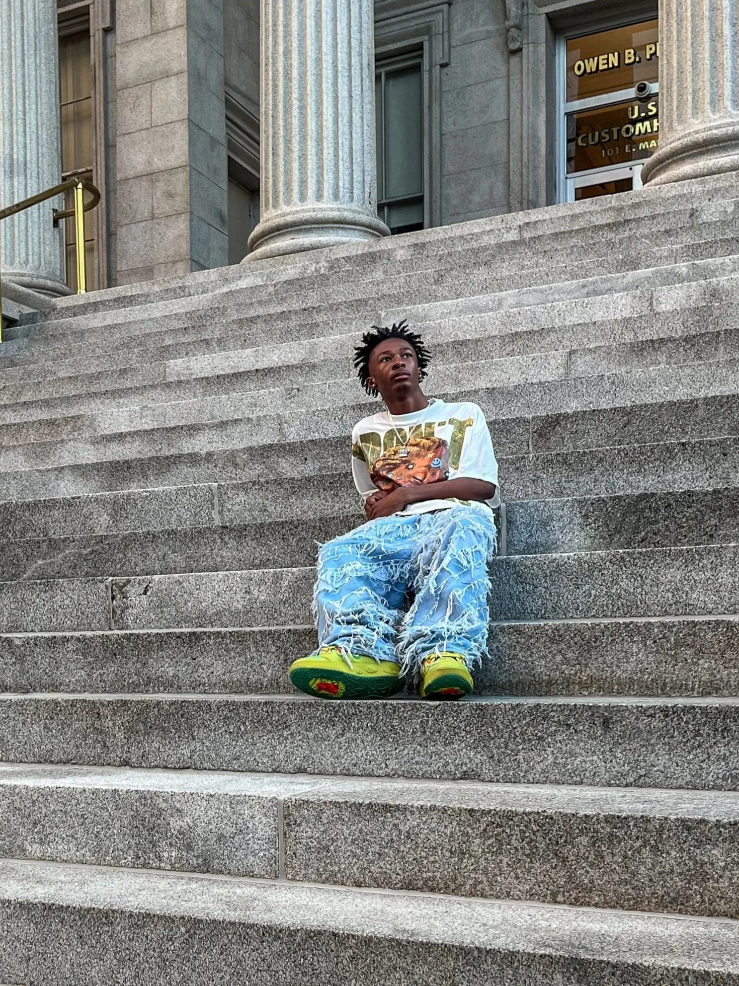 A person with short spiky hair sitting on the steps outside a building with large pillars. The person is wearing a white graphic t-shirt, distressed blue jeans, and bright yellow shoes.