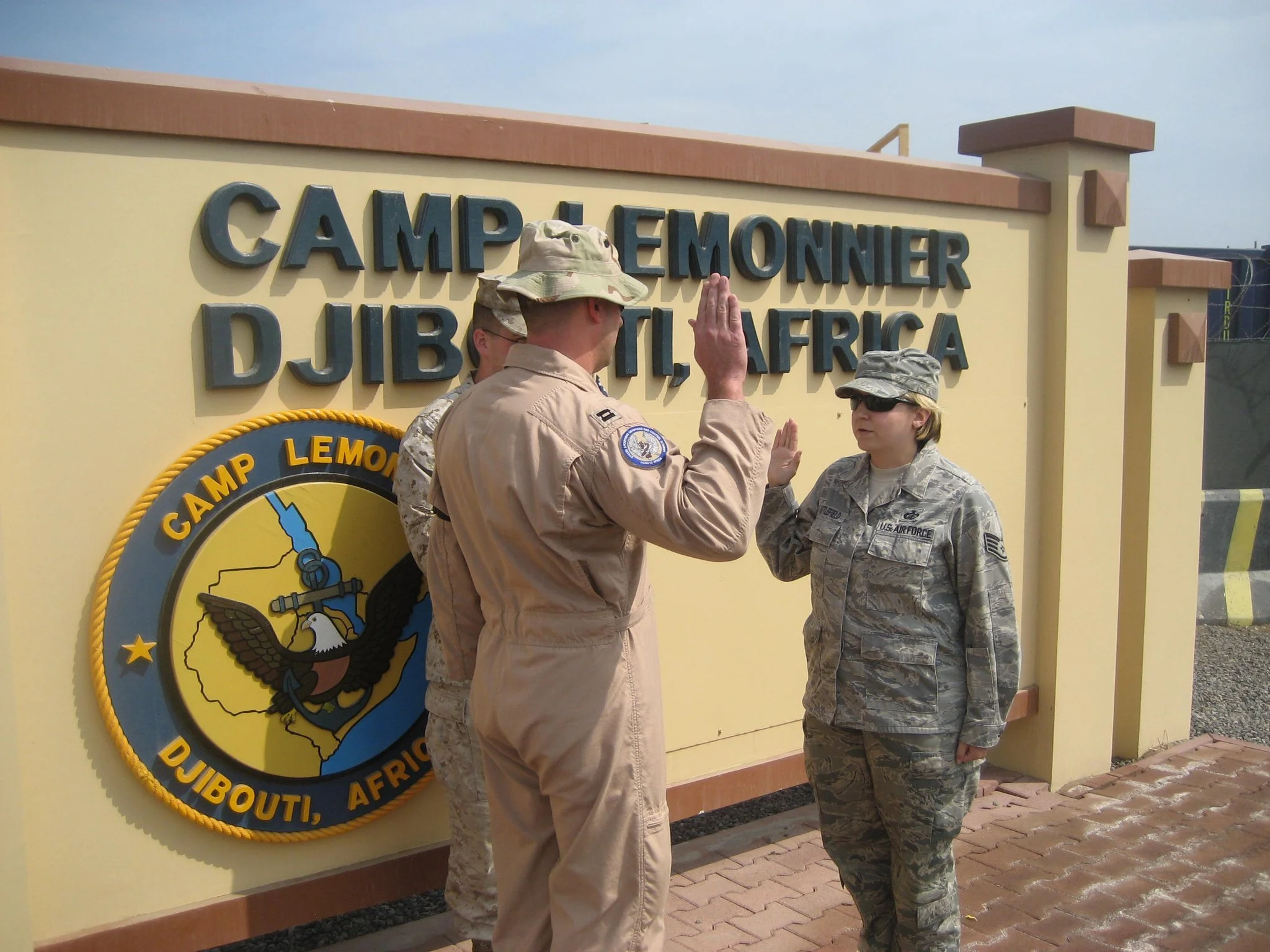 Two military personnel, one male and one female, giving each other a high five in front of a yellow sign that reads "Camp Lemonnier Djibouti, Africa". The male is in a tan uniform and the female in a camouflage uniform, both wearing hats and sunglasses.