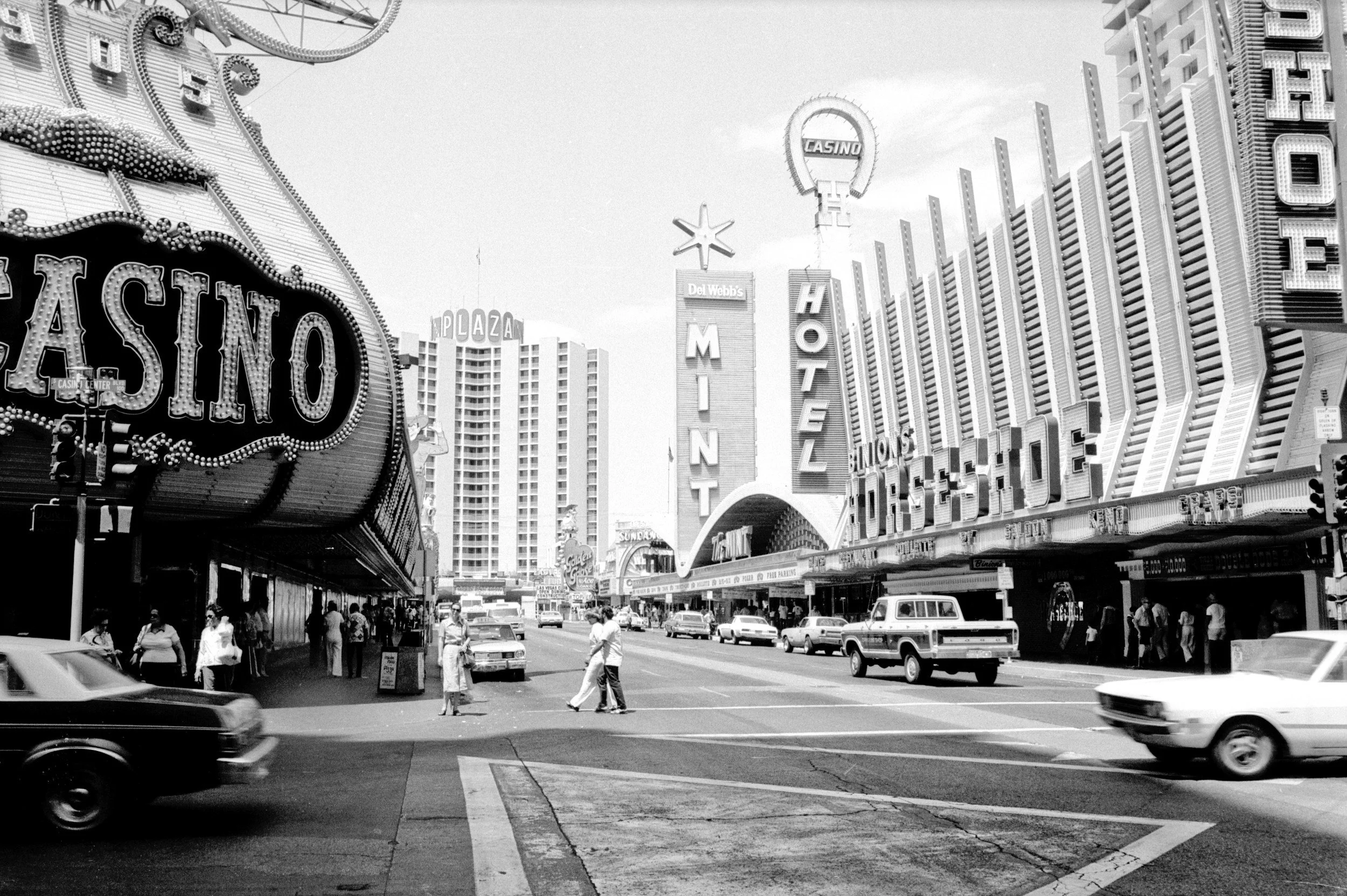 Black and white photo of a busy city street with casino signs, including a large 'CASINO' sign, tall hotel buildings, pedestrians crossing the street, and vintage cars driving by.