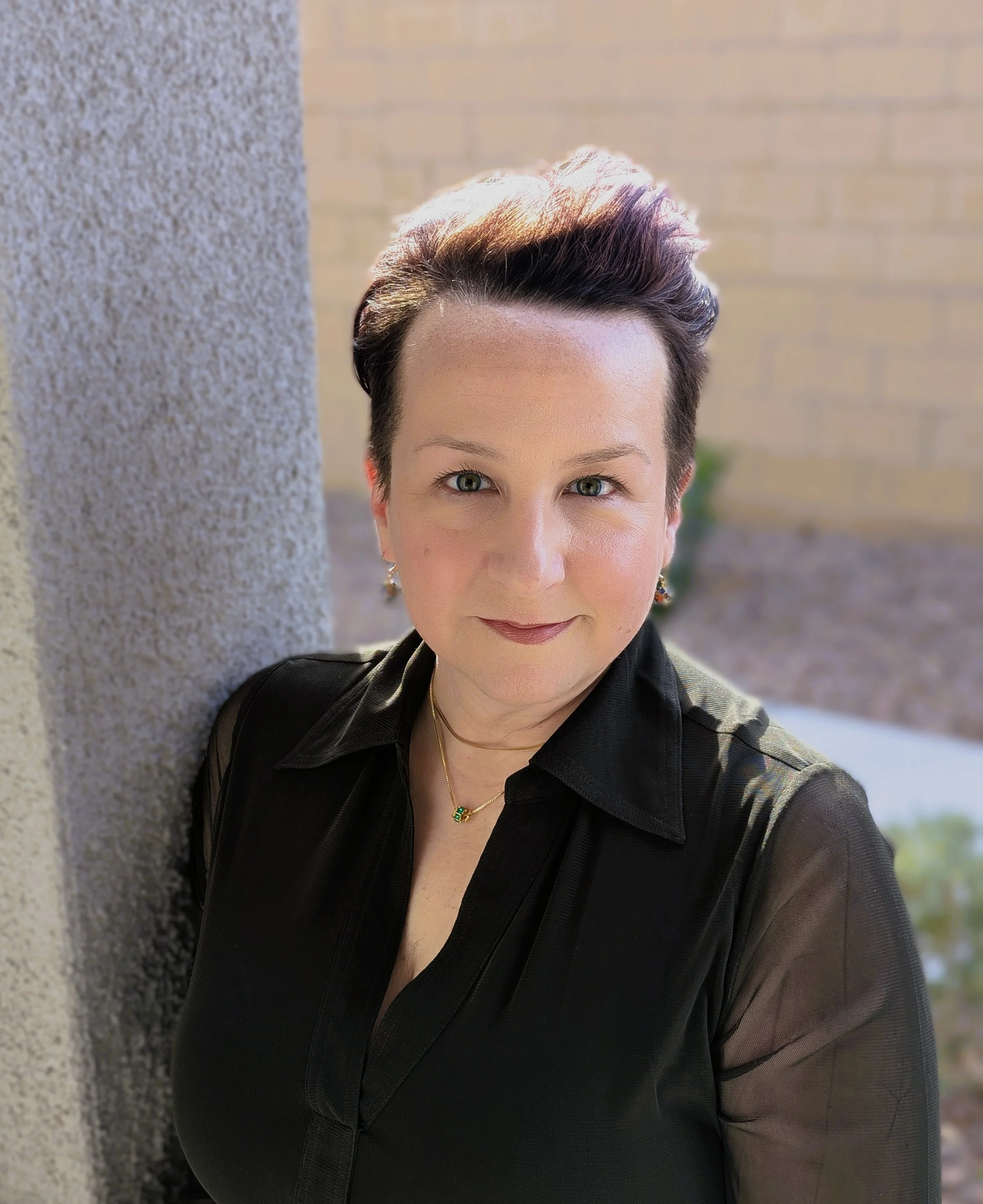A woman with short dark hair, wearing a black top and jewelry, standing outdoors by a textured wall with a blurred background of a brick wall and trees.