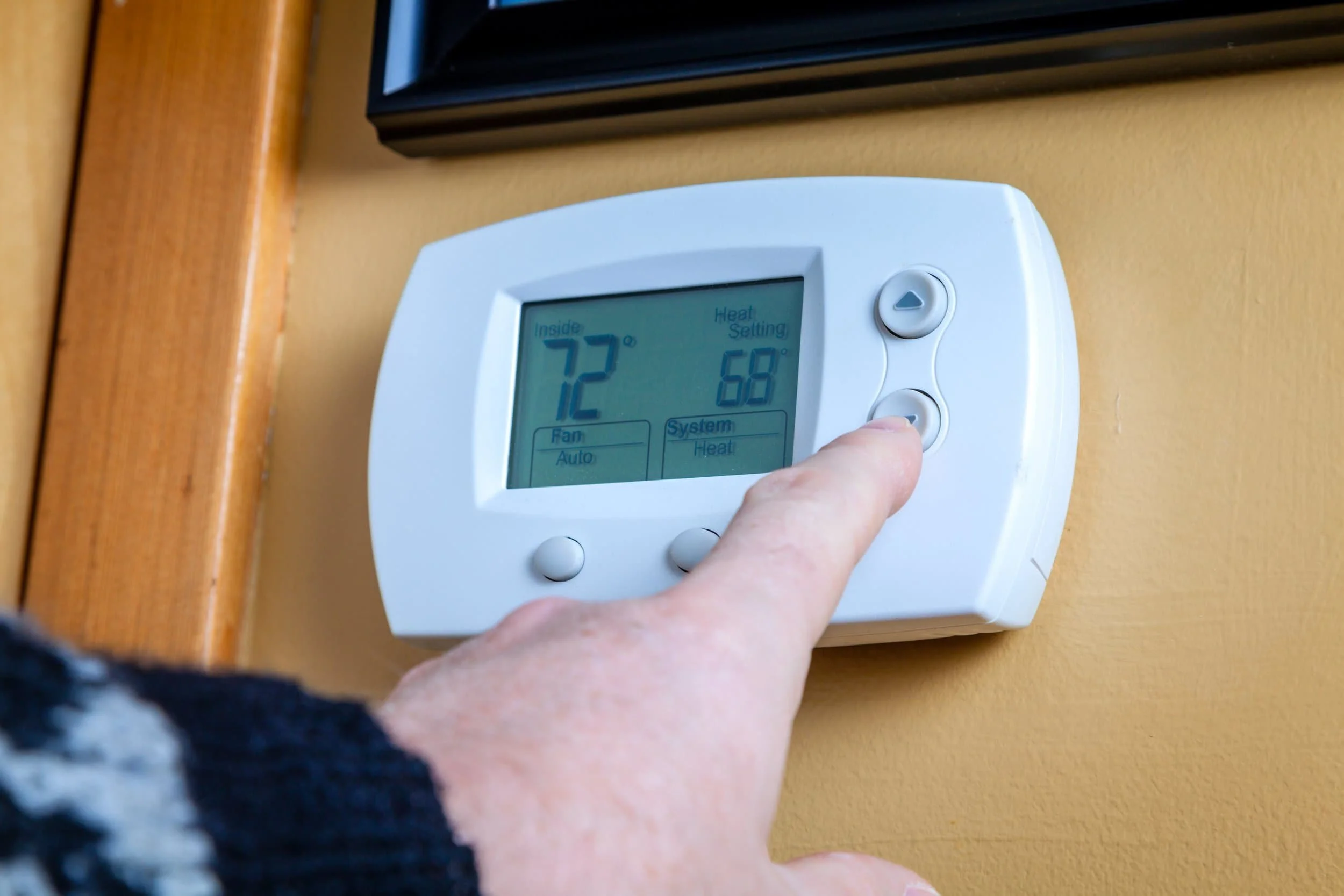 A hand adjusting a digital thermostat set to 72°F inside, with a heat setting of 68°F, mounted on a yellow wall.