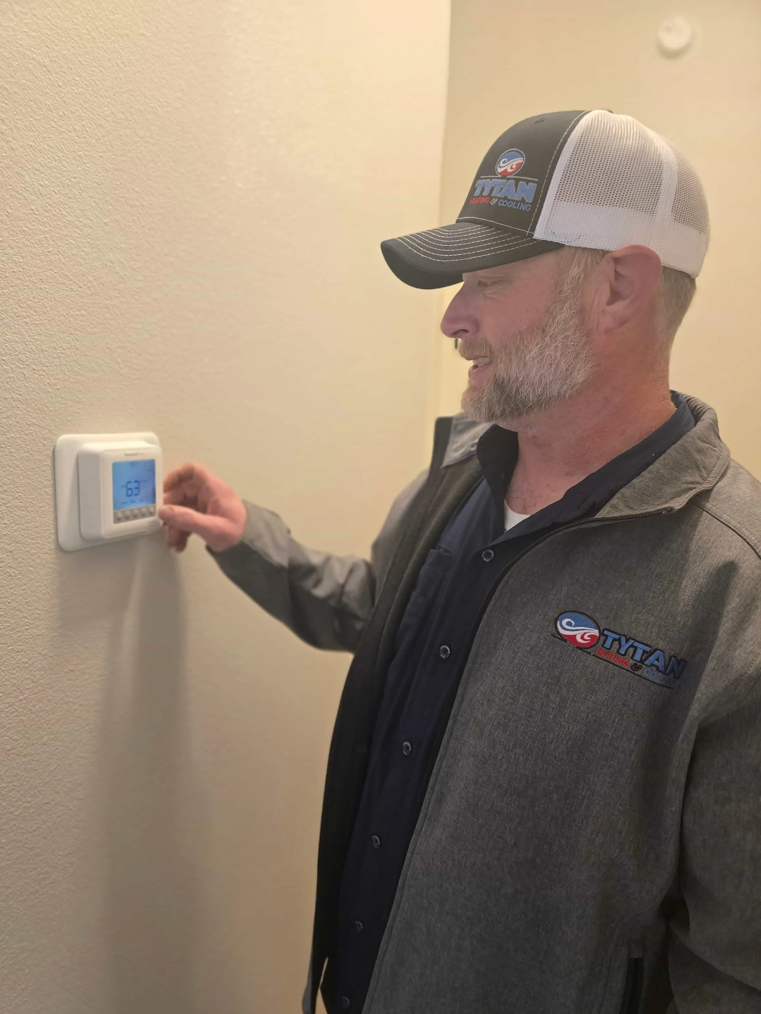 Man in a Titan uniform adjusts a wall thermostat displaying 63°F.