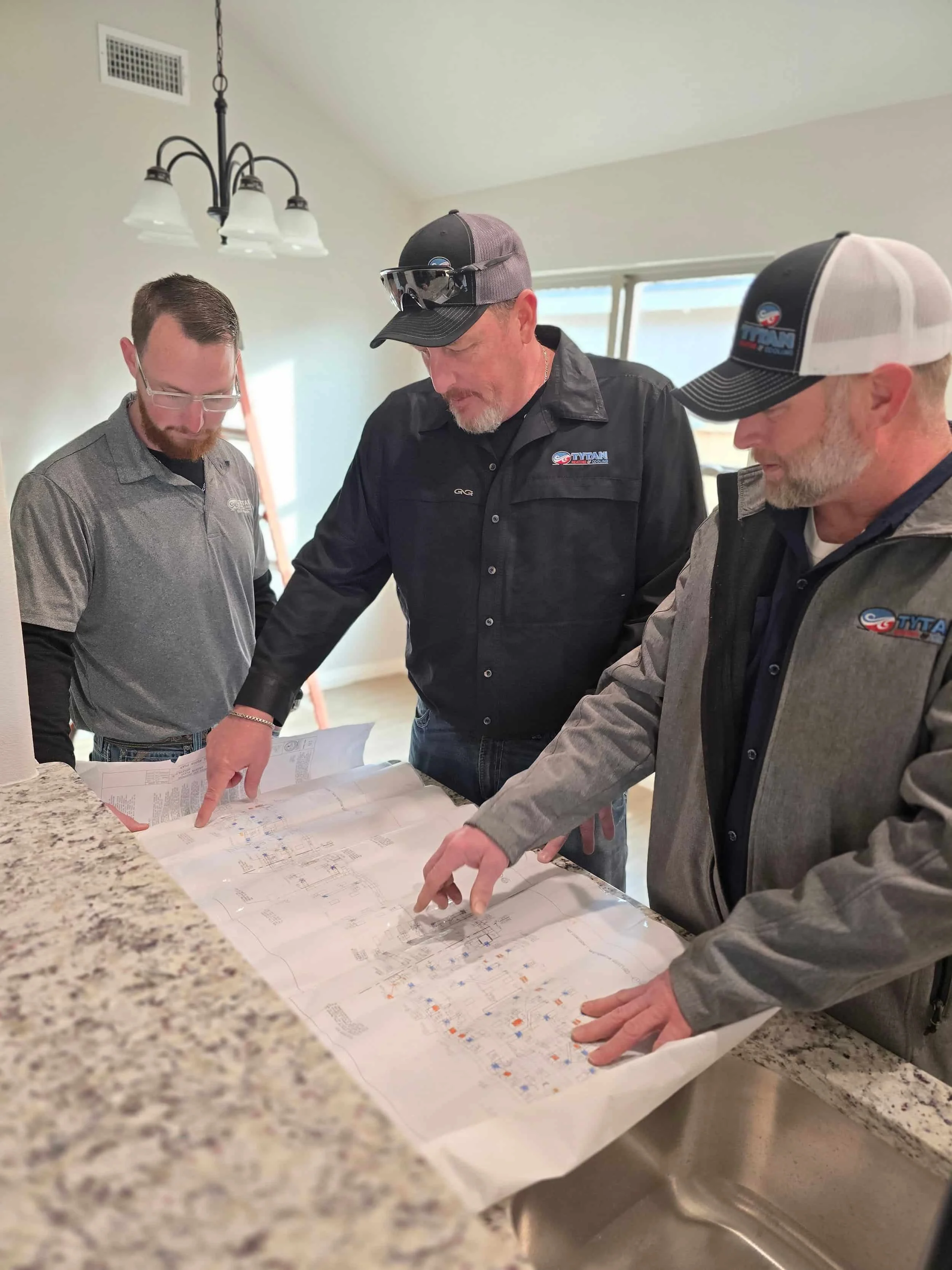 Three men in work uniforms discuss blueprints spread out on a kitchen counter indoors.