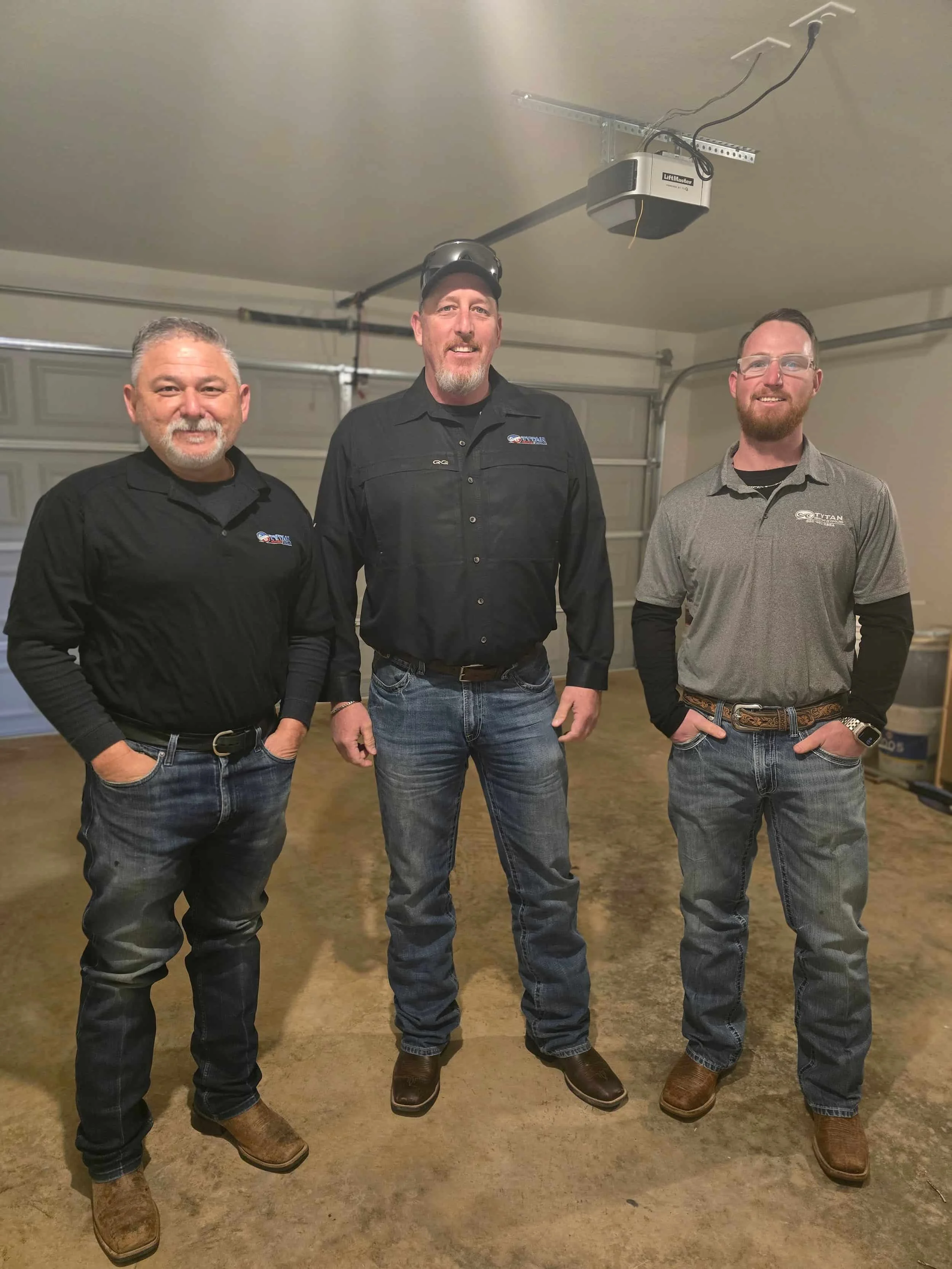 Three men in casual work shirts and jeans stand side by side in a spacious garage, smiling at the camera.