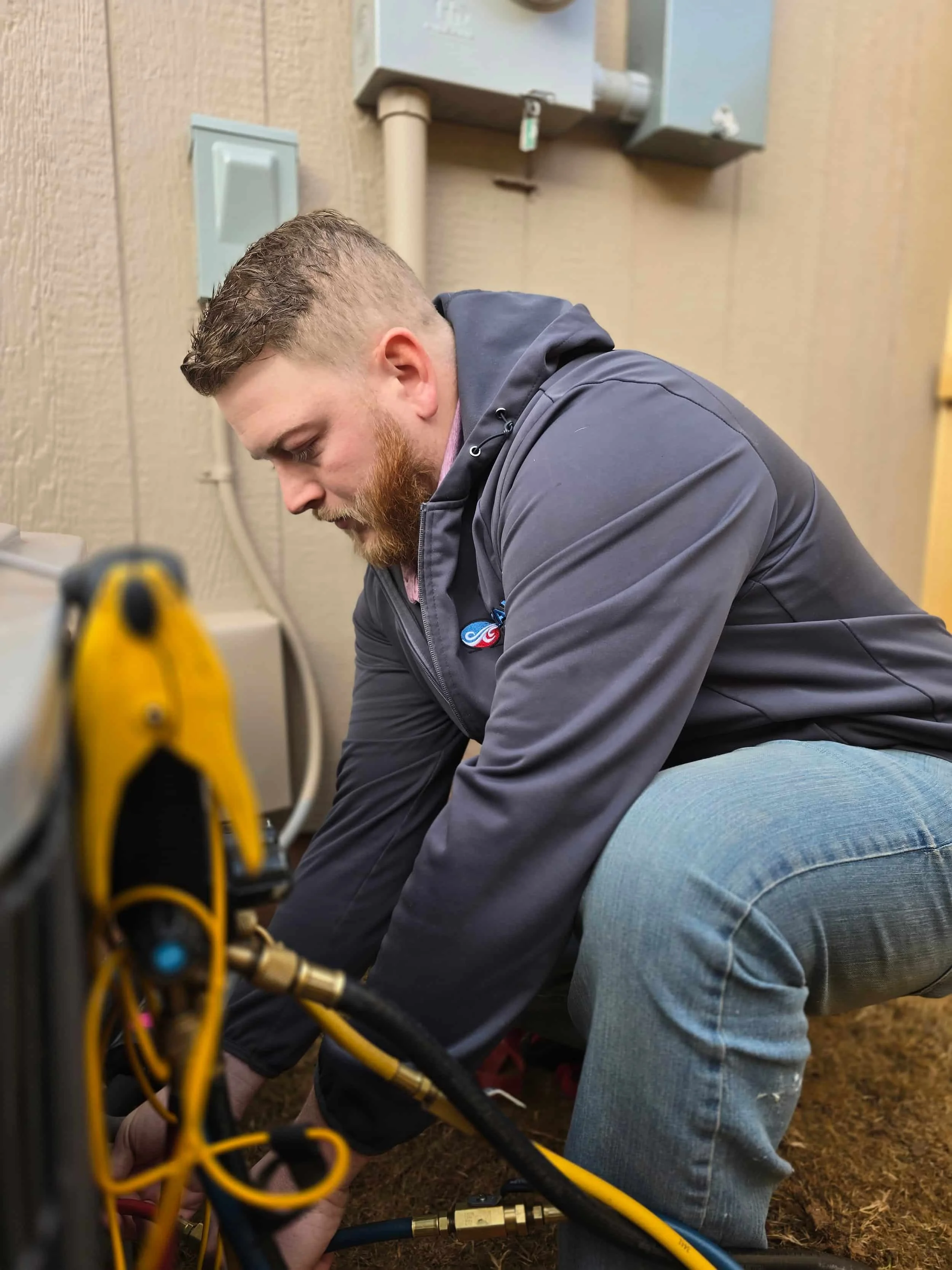 A technician kneels outside, working on HVAC equipment next to a building wall, wearing a gray hoodie and jeans.