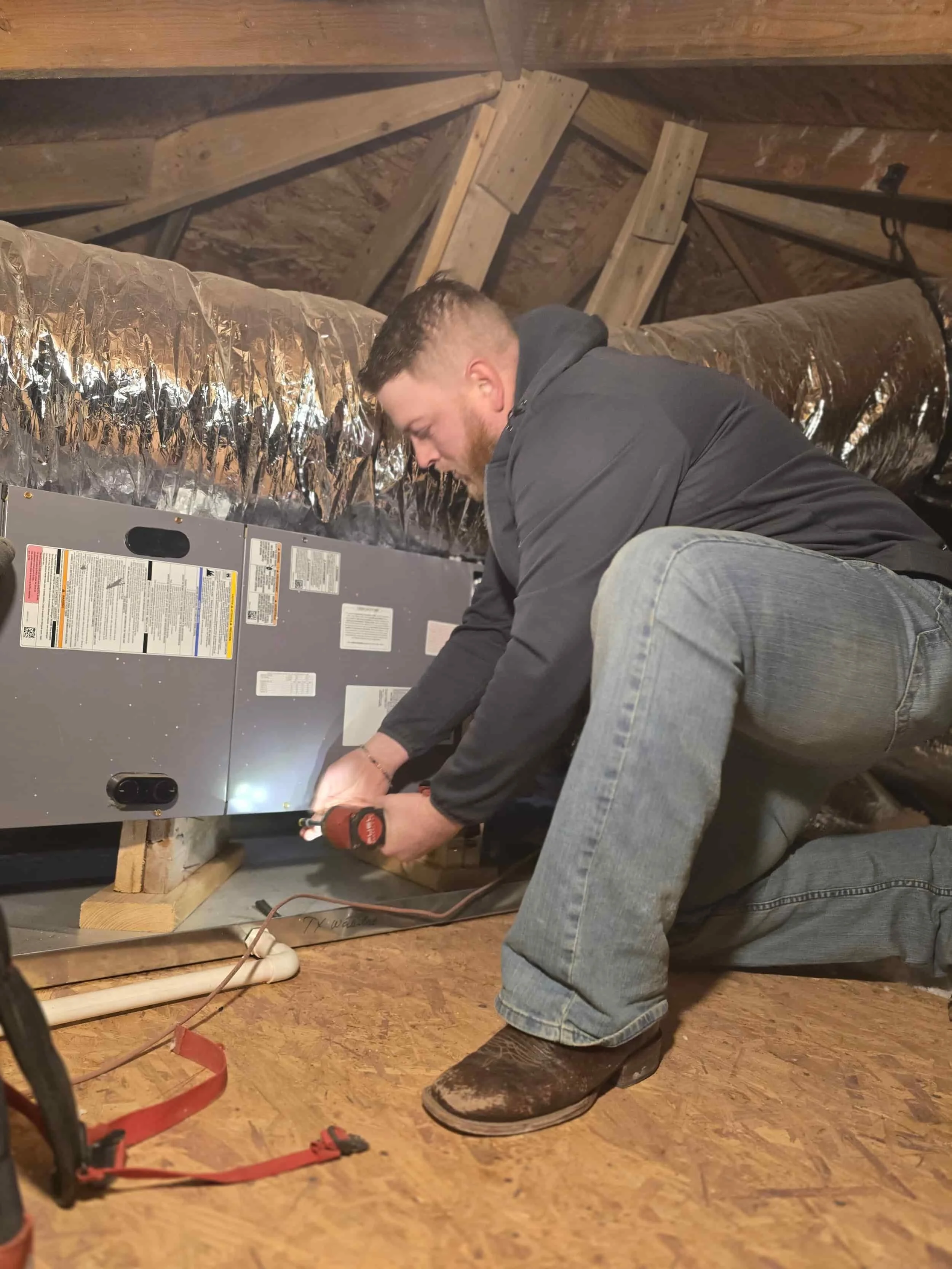 A man kneels in an attic, using a power drill to work on HVAC equipment, with ducts and exposed wooden beams visible.