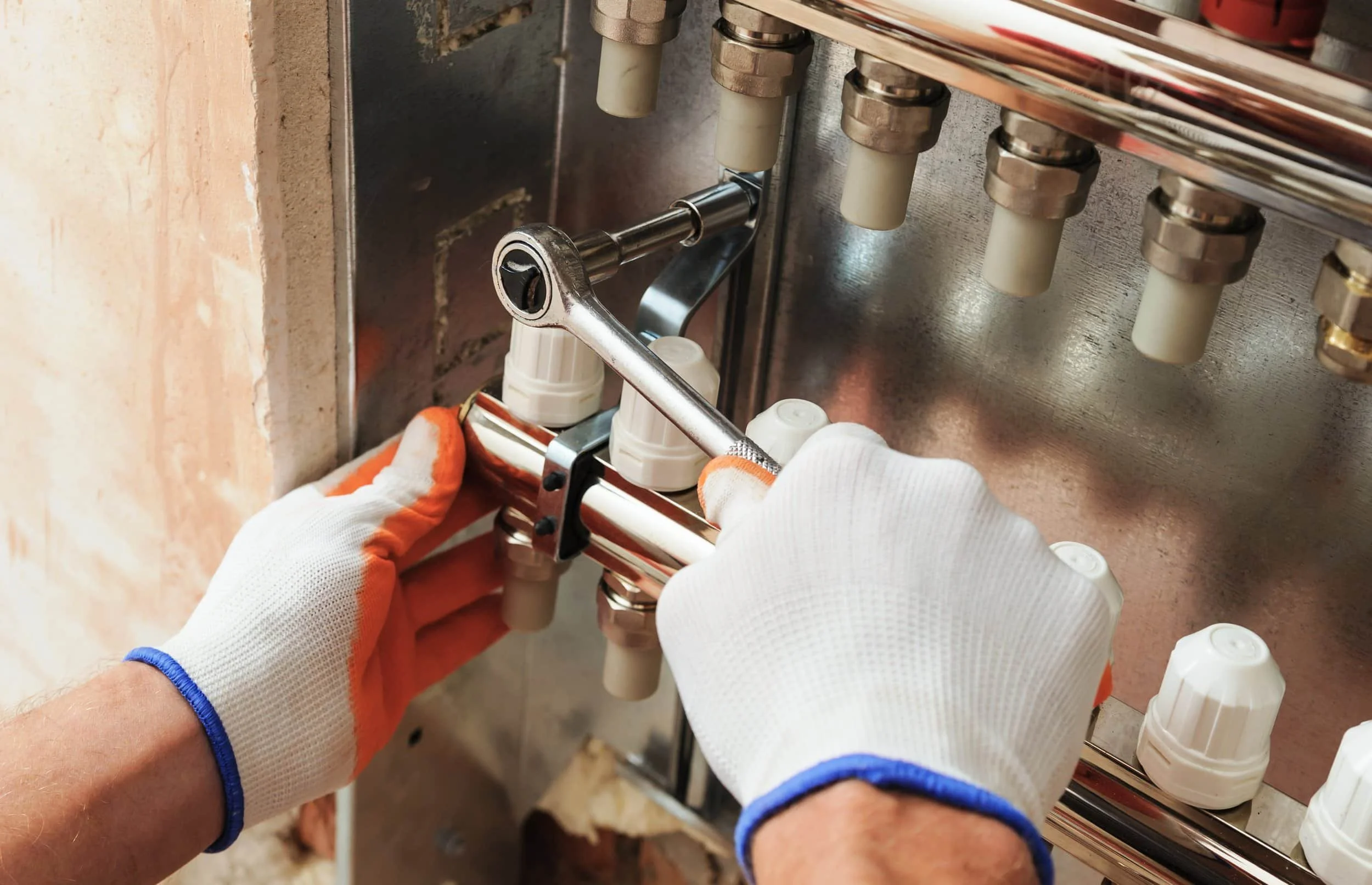 A person wearing gloves uses a wrench to tighten plumbing connections on a metal pipe manifold.