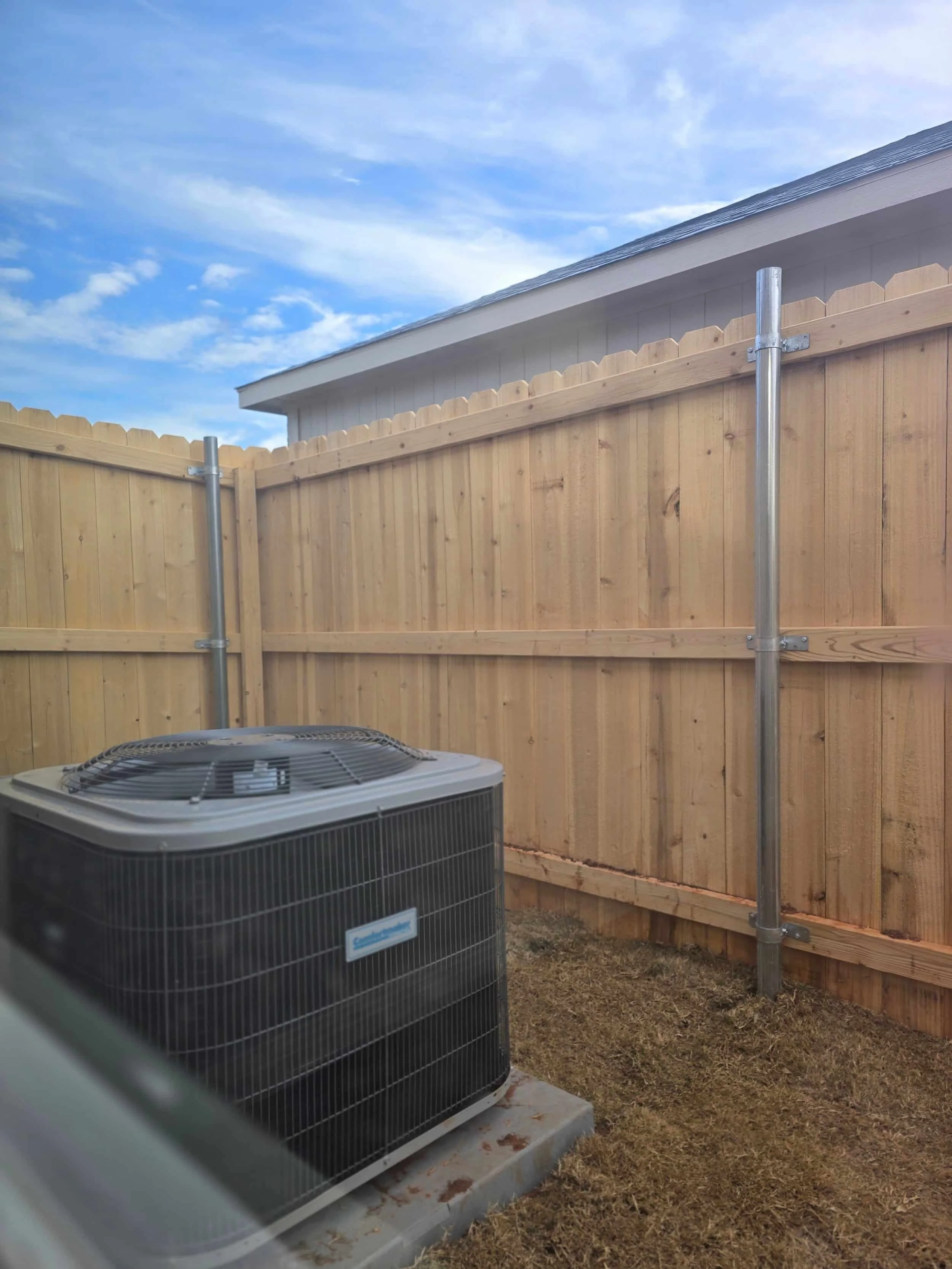 A central air conditioning unit sits on dry grass near a wooden fence and storage shed under a party cloudy sky.