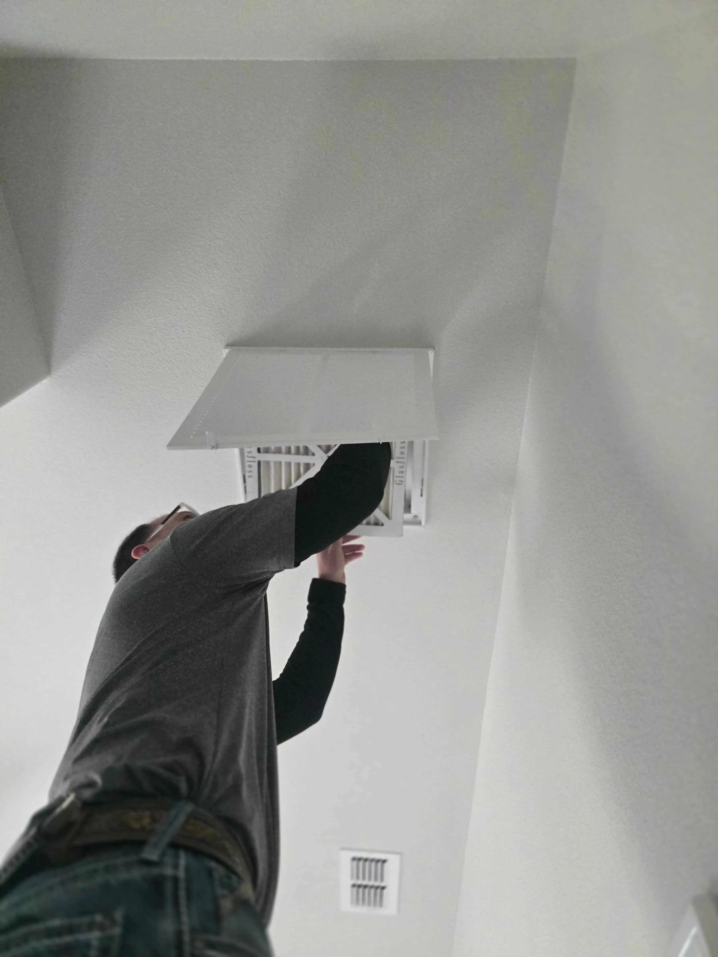 A person in a gray shirt stands on a ladder, changing or fixing an air vent on a white ceiling