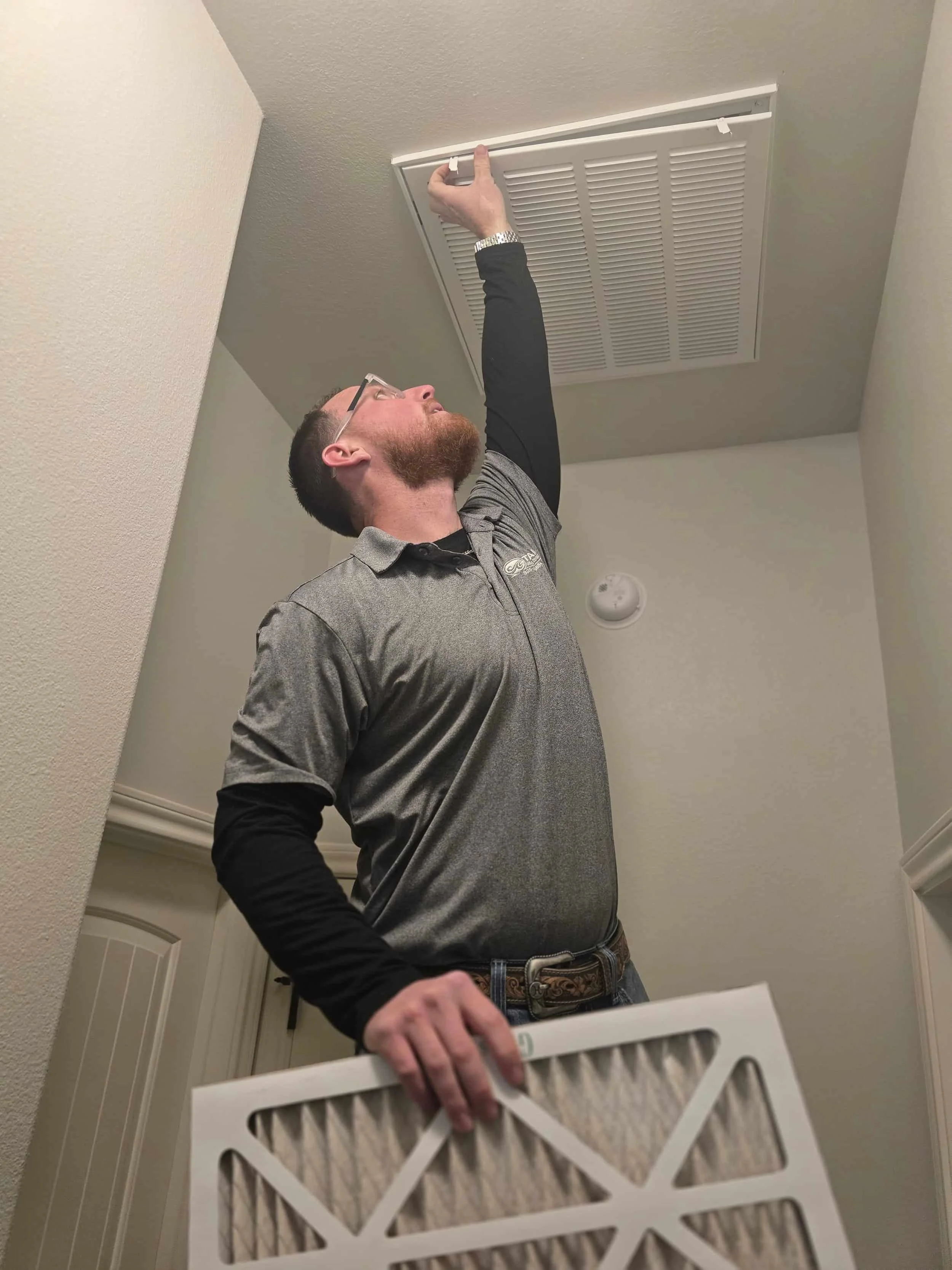 Man changing an air filter in a ceiling vent, standing on the floor and reaching up with a new filter in hand.