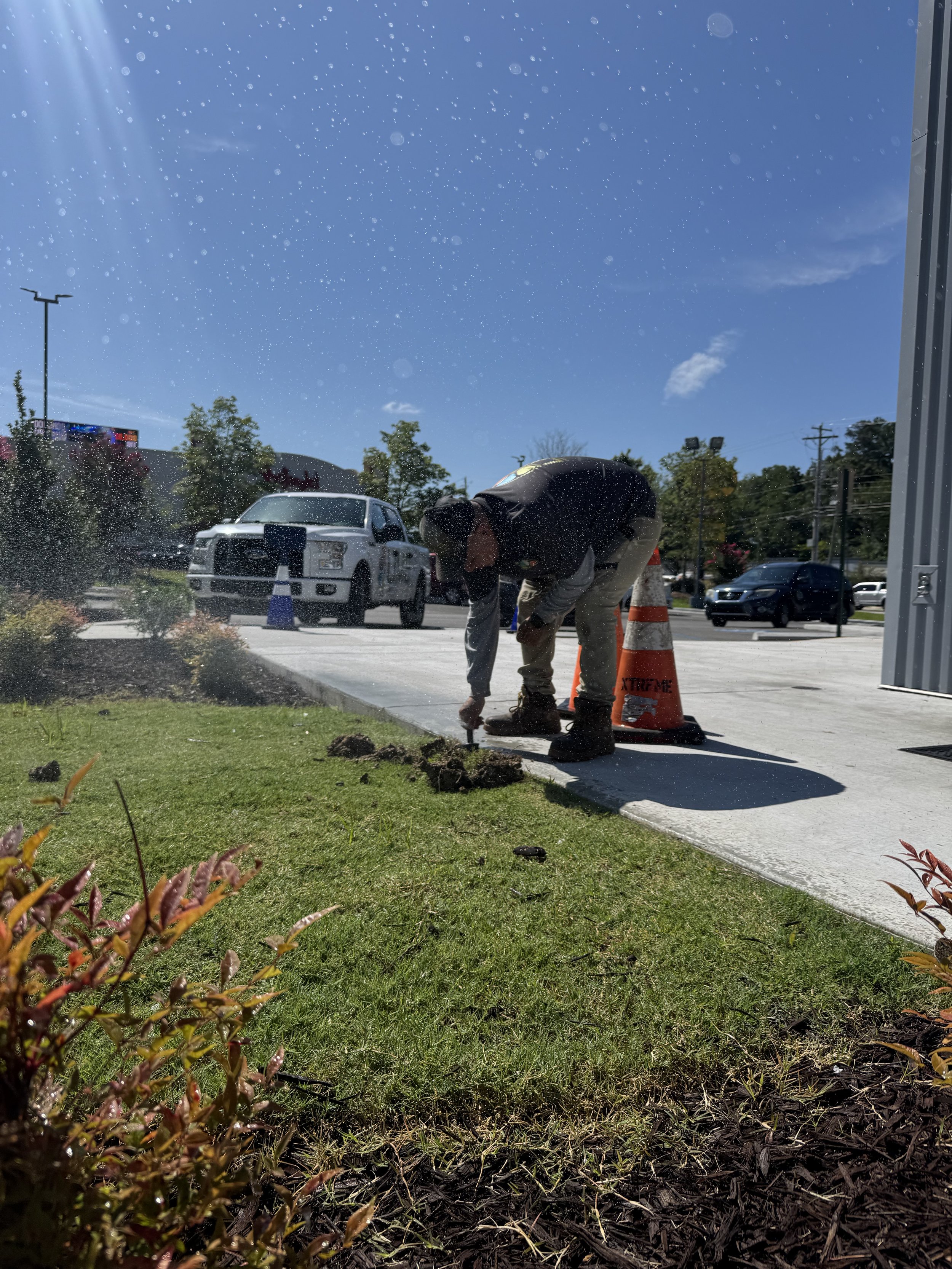 A LIME worker is repairing or inspecting the irrigation near a sidewalk, kneeling with a tool in hand as water sprays around him. Traffic cones and a white truck sit nearby under a bright, sunny sky.