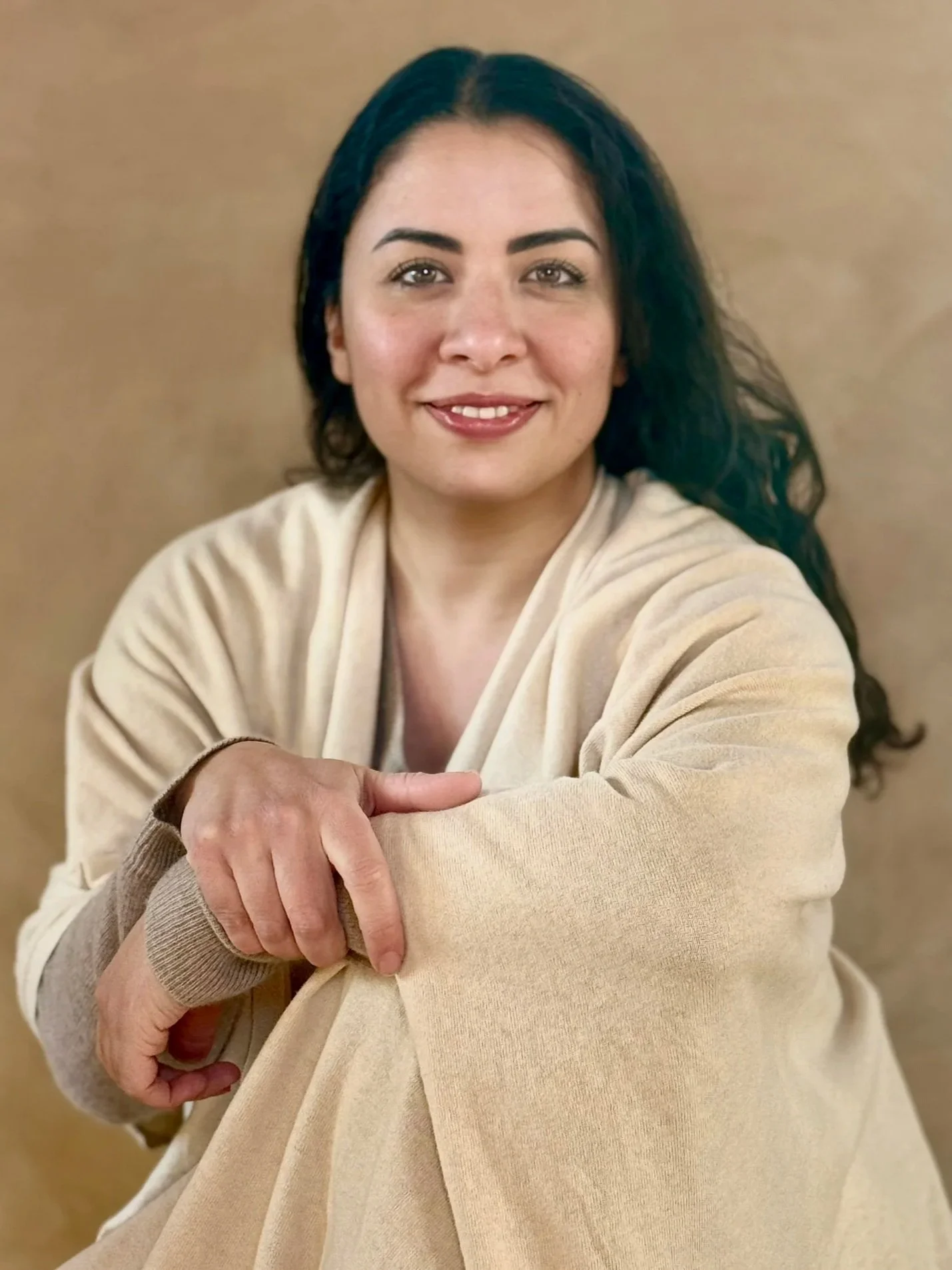 A woman with dark wavy hair, wearing a beige sweater, smiling and looking at the camera against a neutral background.