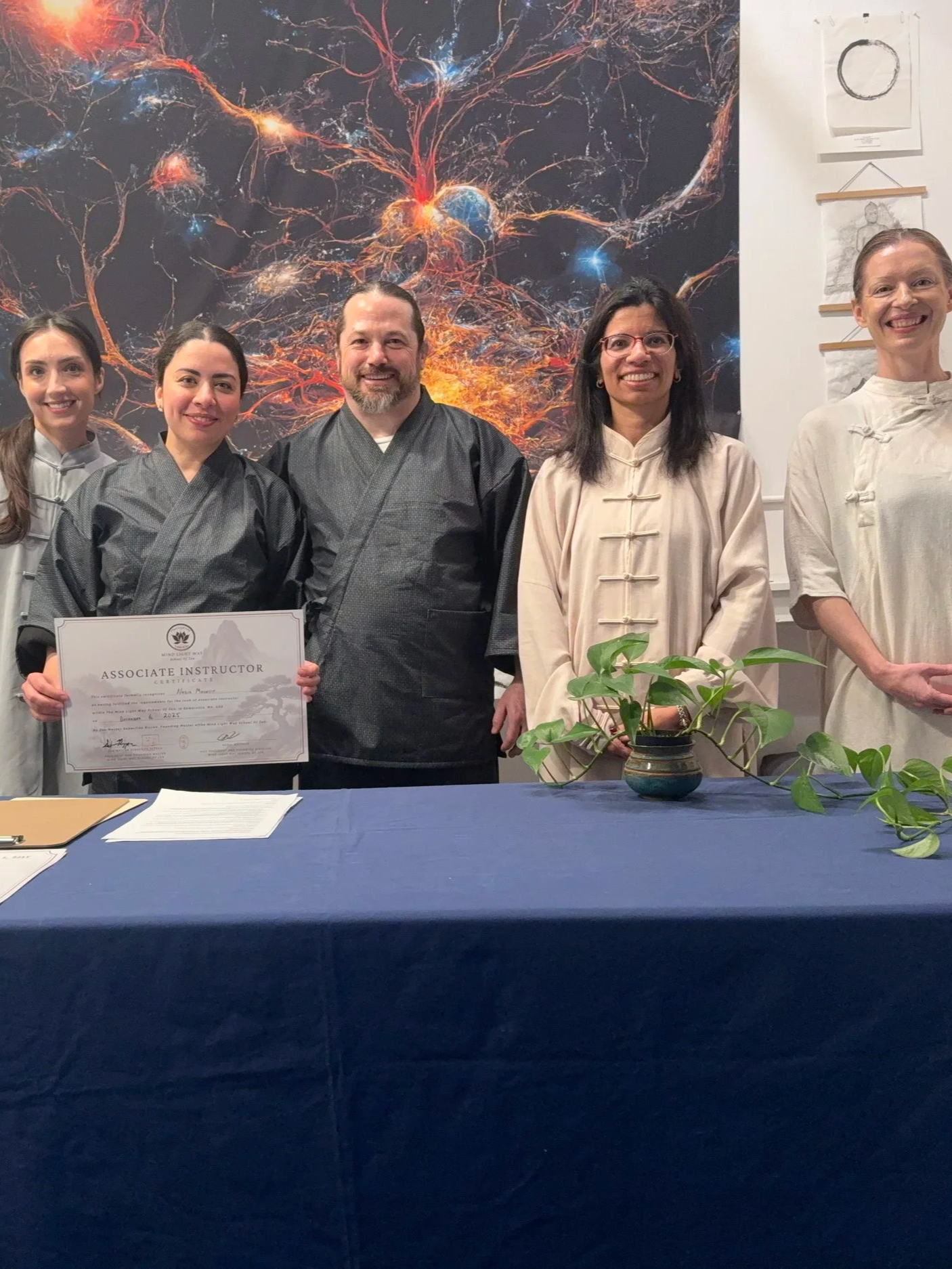 Group of five people in traditional Asian attire standing behind a table, with one person holding an 'Associate Instructor' certificate. There is a large, colorful artwork of neural networks or cosmic scene in the background and a potted plant on the table.