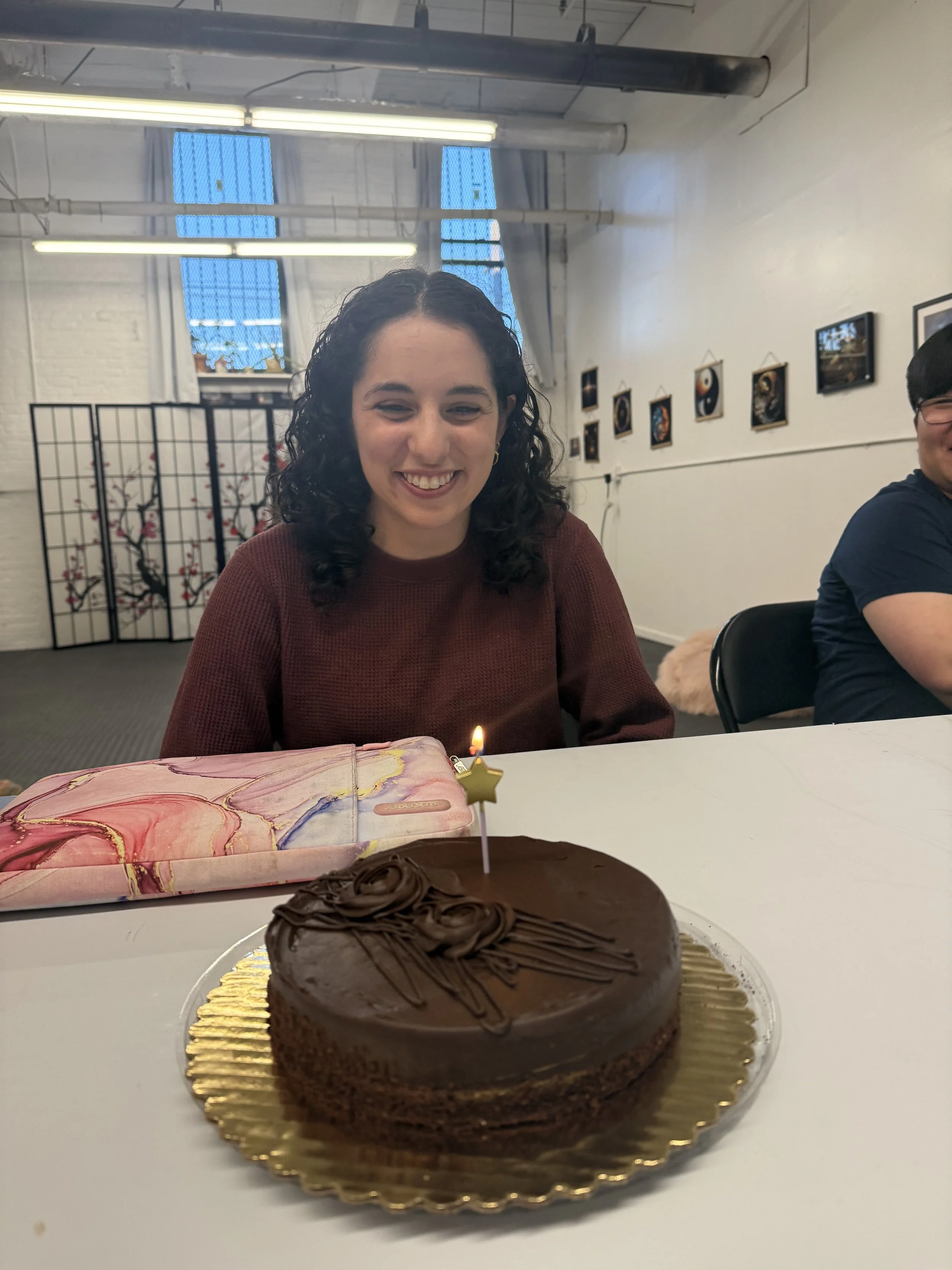 A young woman with curly dark hair smiling at a birthday celebration, sitting behind a chocolate cake with a lit candle, in an indoor space with framed photos on the wall.
