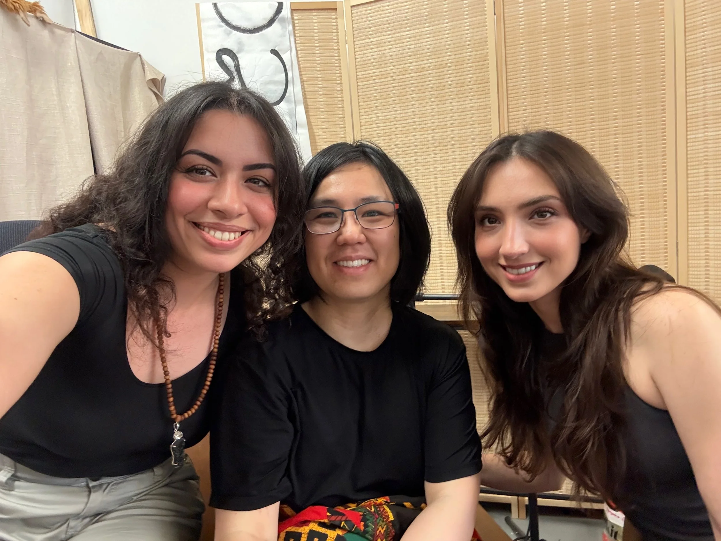 Three young women smiling and taking a selfie together indoors, with light-colored walls and a beige curtain in the background.