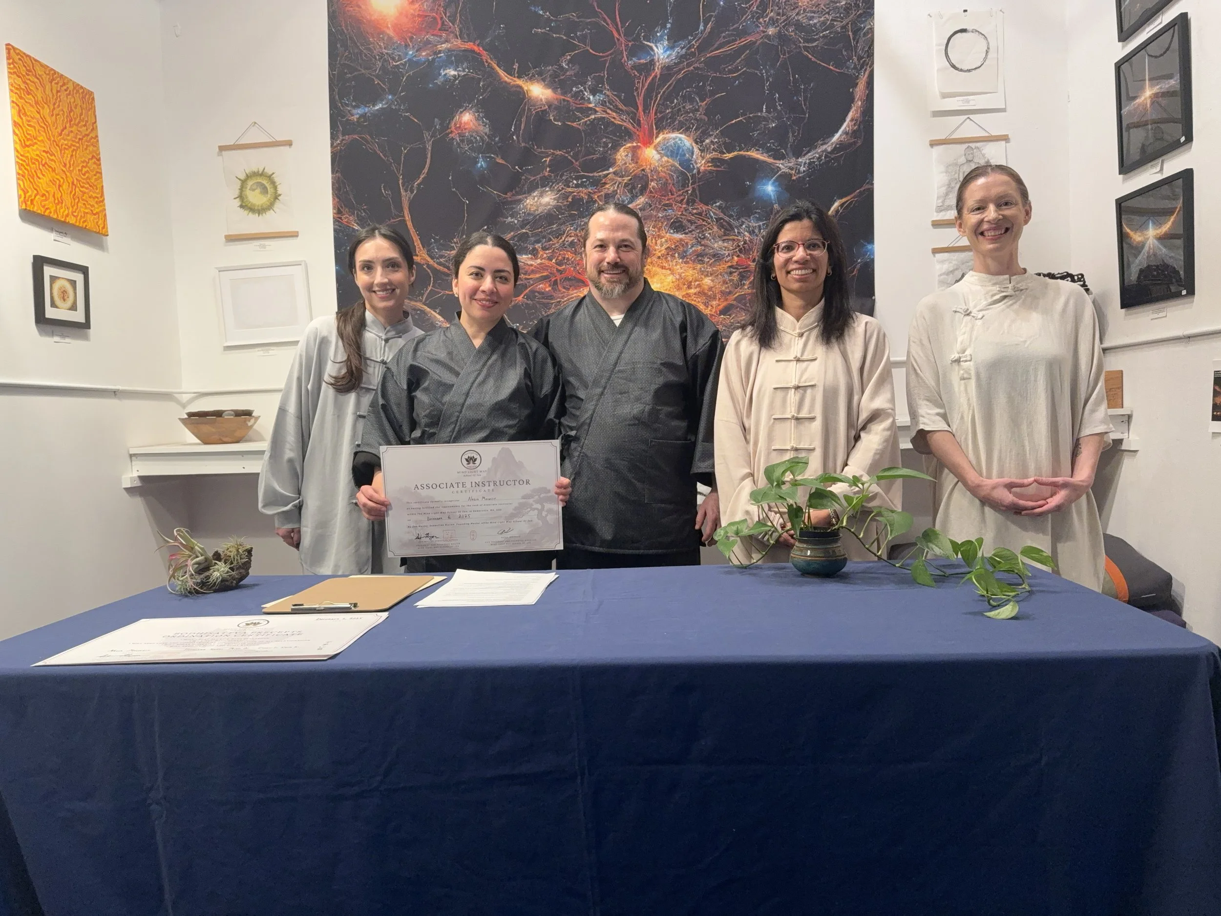 Five people in traditional Asian attire standing behind a table with a blue cloth, with one woman holding a certificate that says 'Associate Instructor.' The background features a large, colorful abstract mural resembling cosmic or neural imagery. The room has framed artwork on the walls and small potted plants on the table.