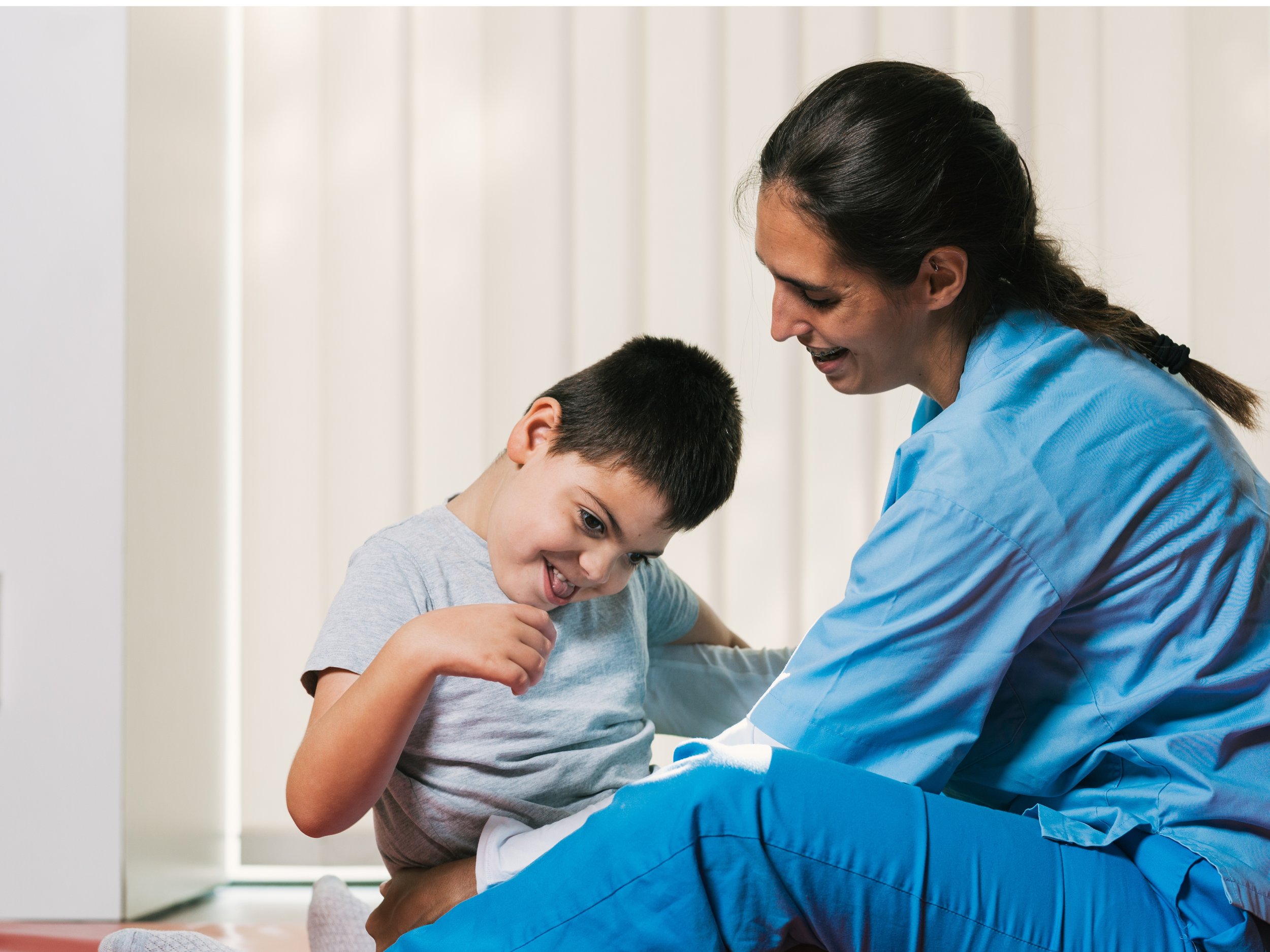 A healthcare worker, dressed in blue medical scrubs, gently examines a young boy who is smiling and laughing during a medical check-up.