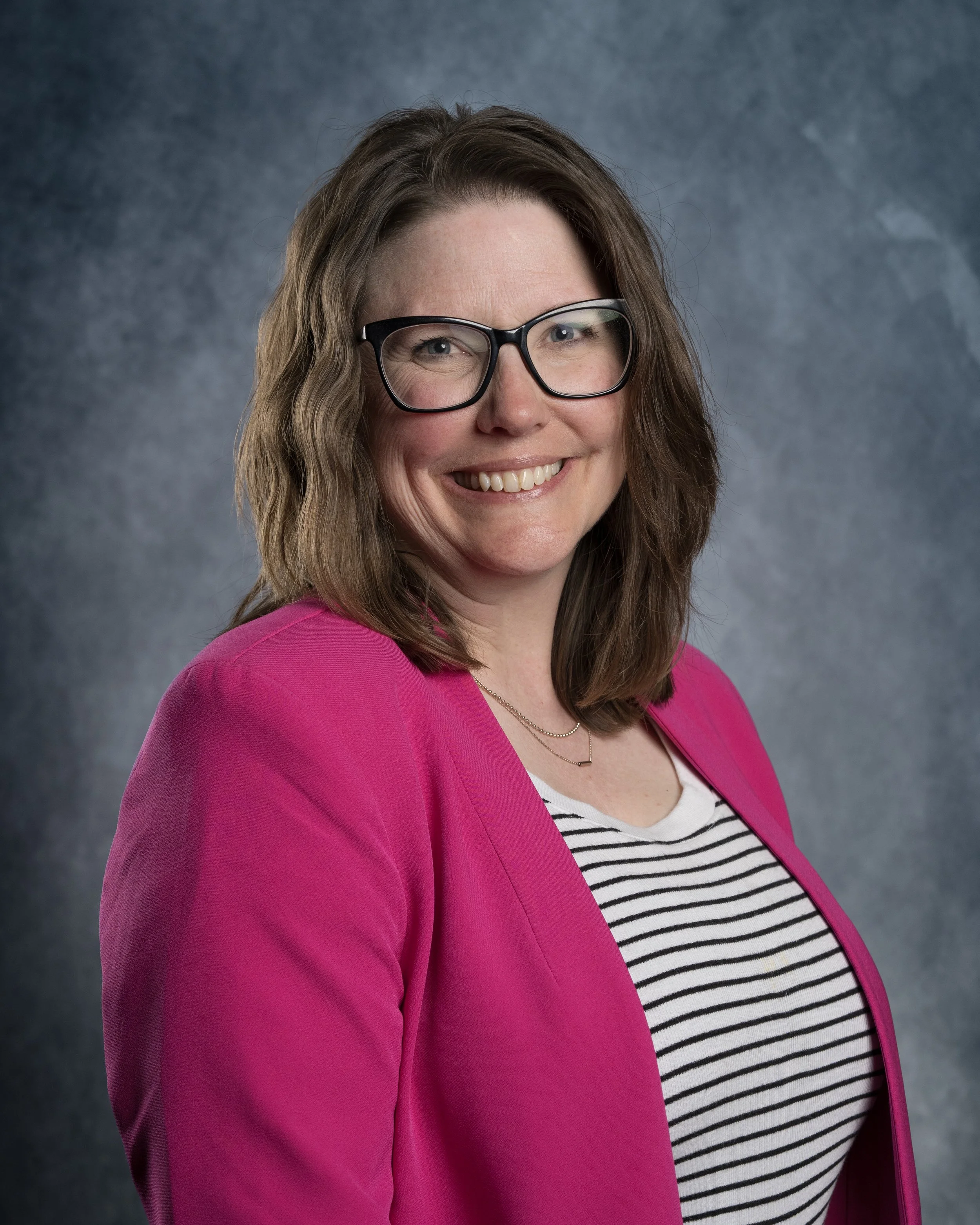 A woman wearing glasses and a colorful purple and black patterned jacket, smiling against a green background.