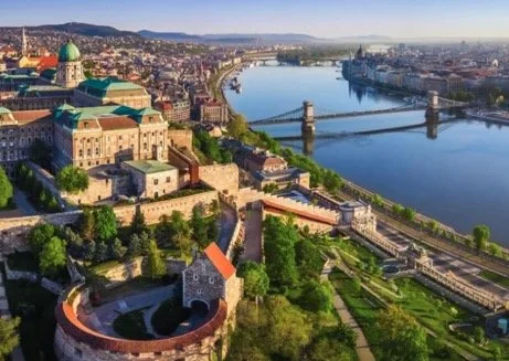 Aerial view of Budapest, Hungary, featuring Buda Castle with its green dome, the Danube River, and the Chain Bridge, surrounded by city buildings and green spaces.